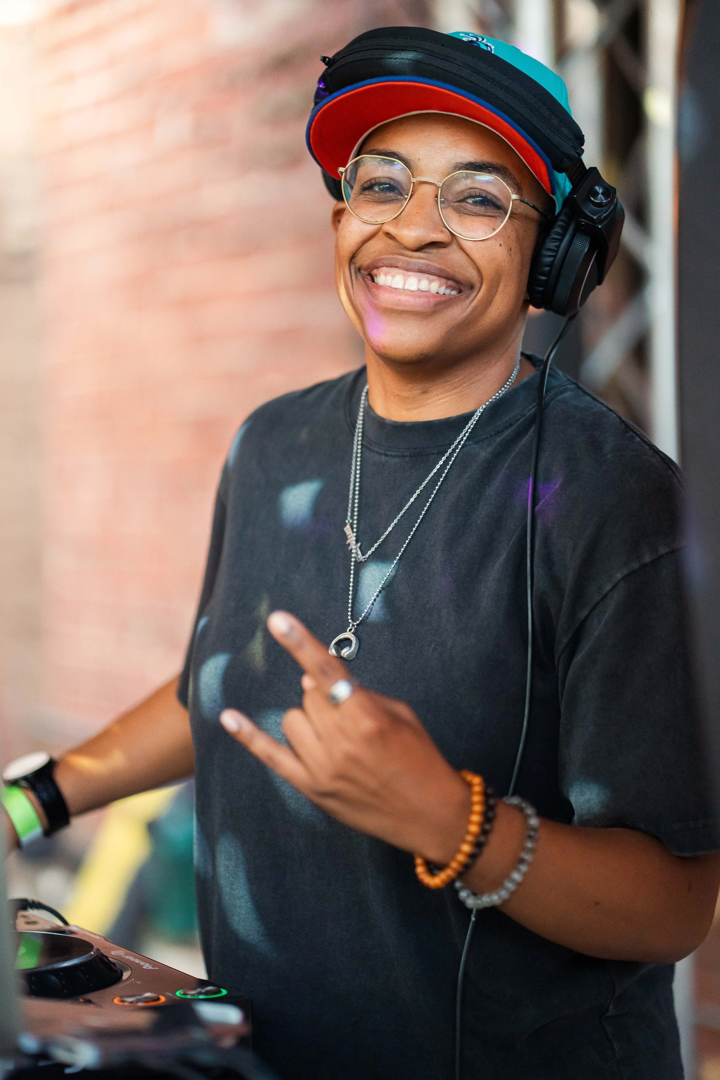 A smiling person wearing glasses, headphones, a colorful hat, and a black t-shirt making a hand sign while standing near a DJ booth.