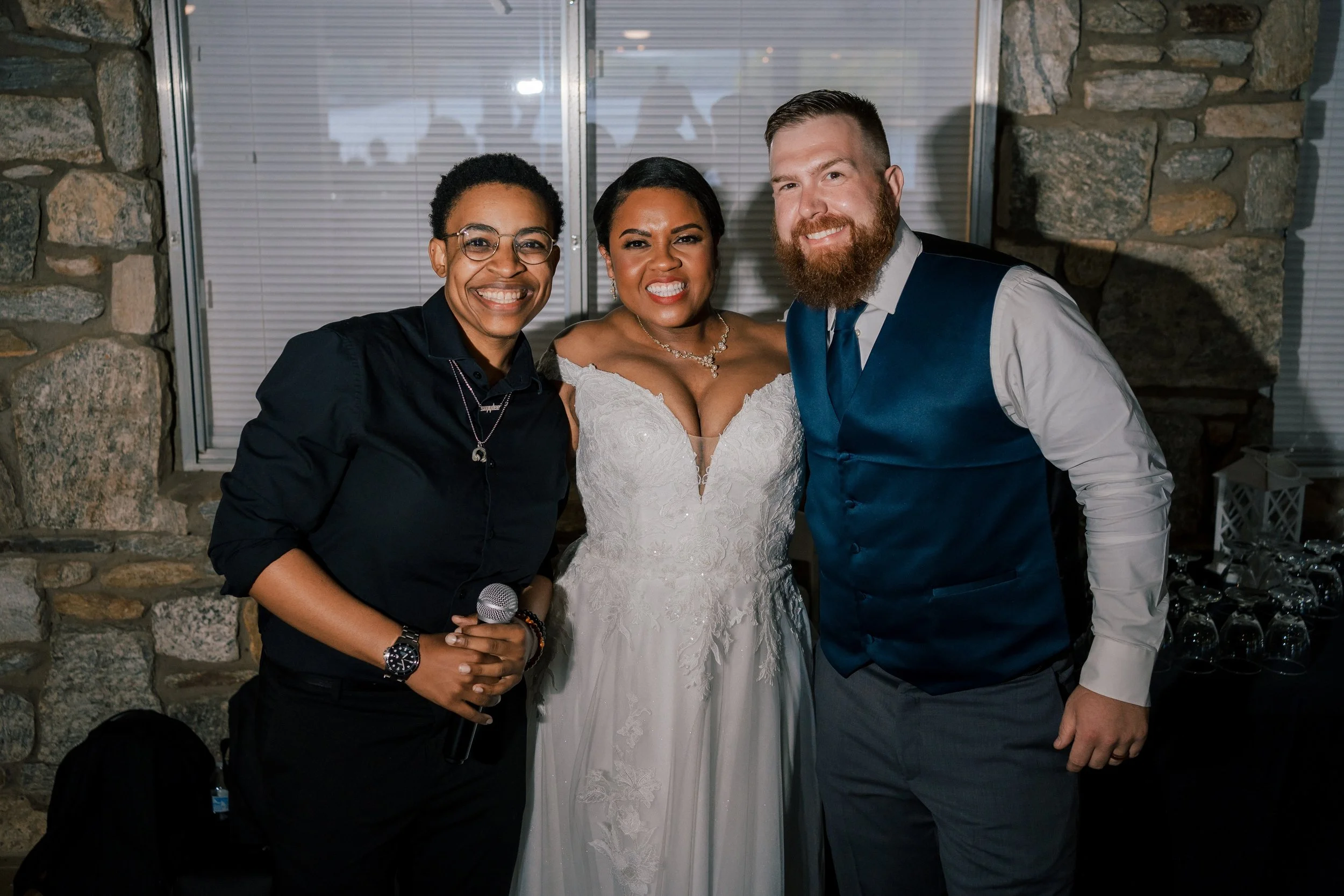 Three people standing together at a wedding reception, with a stone wall and window behind them. The woman in the middle is in a wedding dress, smiling, with a necklace. The woman on the left is holding a microphone, wearing glasses and dark clothing. The man on the right has a beard, wearing a white shirt, blue vest, and gray trousers, smiling.