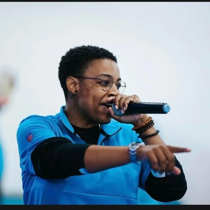 A woman with short curly hair, wearing glasses and a blue top, is holding a microphone and pointing forward, possibly speaking or performing.