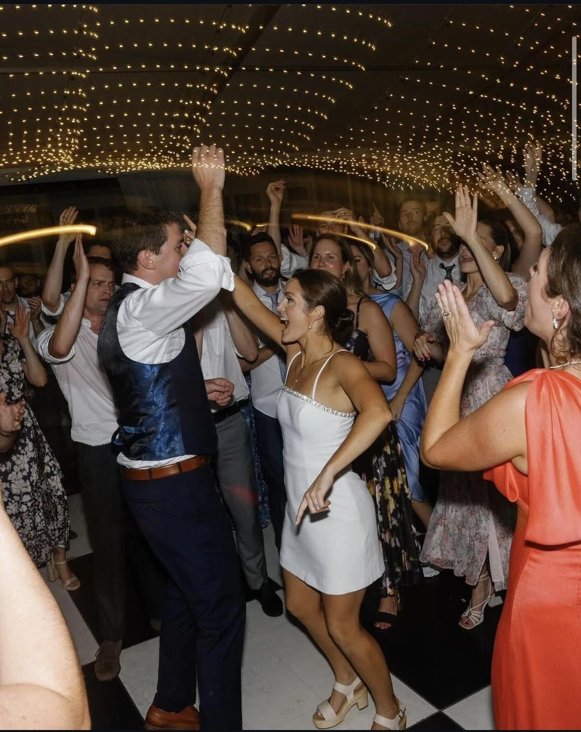 People dancing and celebrating at a wedding reception with string lights overhead.