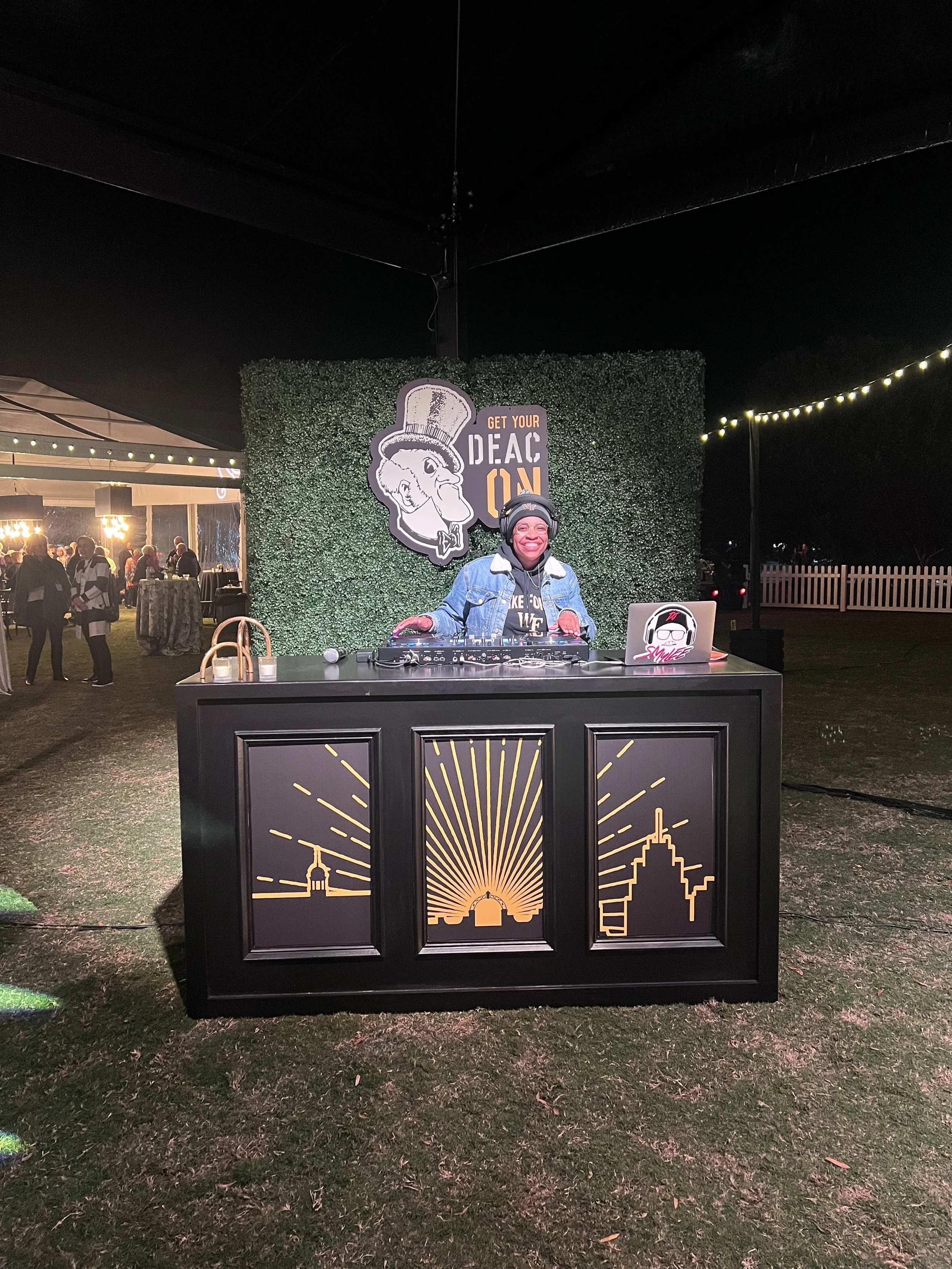 A person DJing at an outdoor event at night, smiling behind a DJ booth with cityscape graphics. The background has a green hedge with signage and strings of lights.