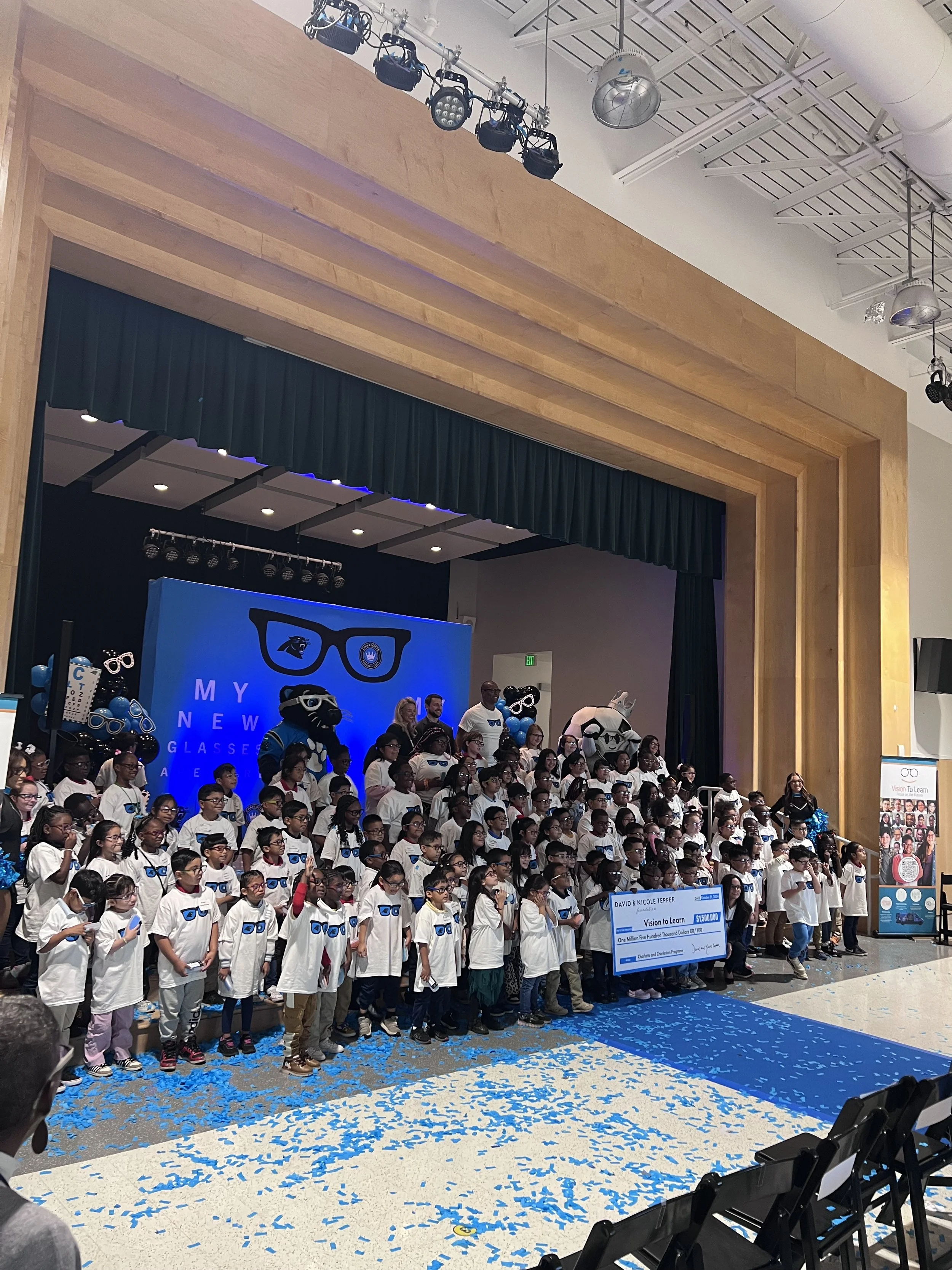 Group of children on stage wearing white T-shirts at a charity event, with mascots, a large blue check, and a blue-themed backdrop.