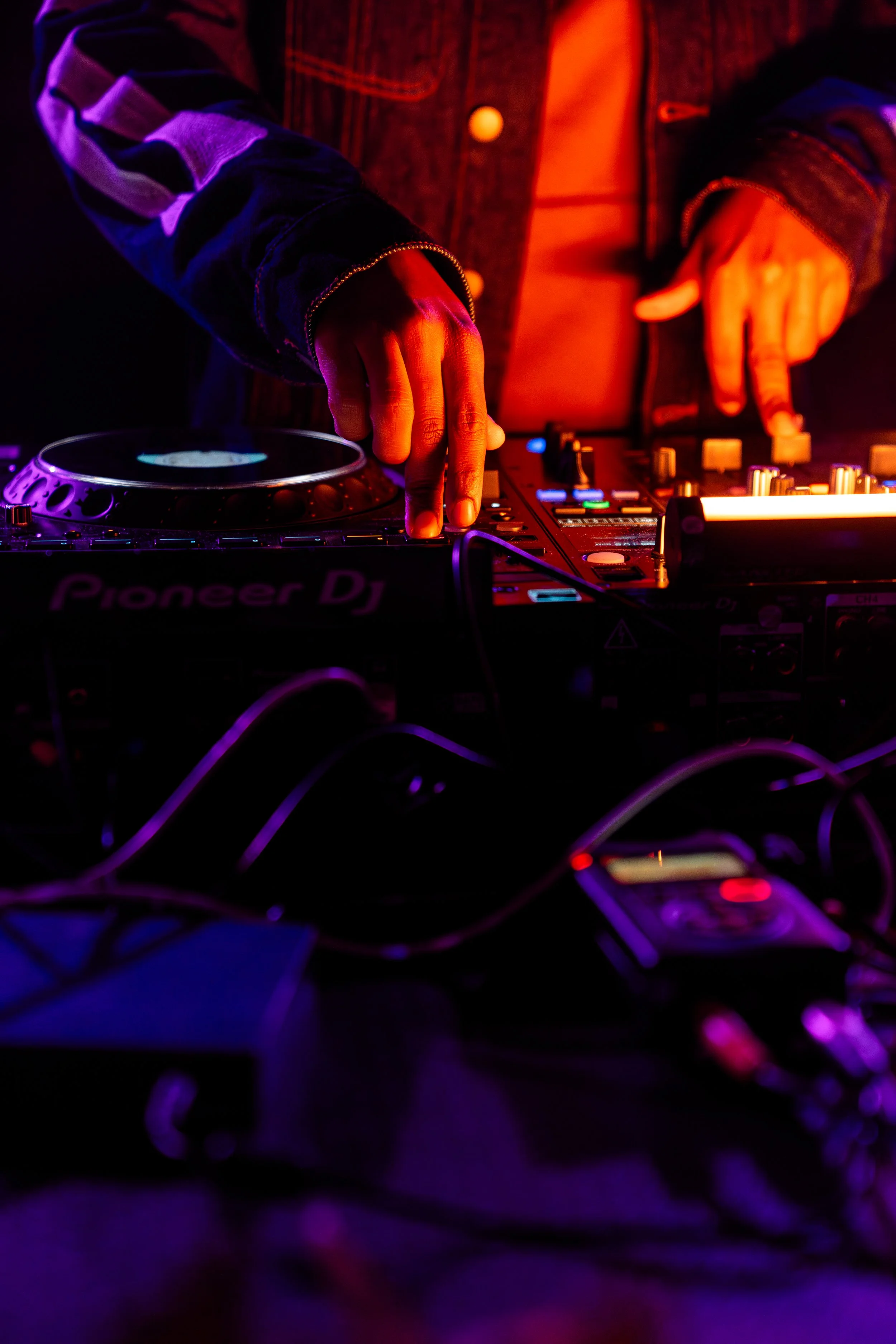 Close-up of a DJ's hands operating a Pioneer DJ controller in a dark setting with colorful stage lighting.