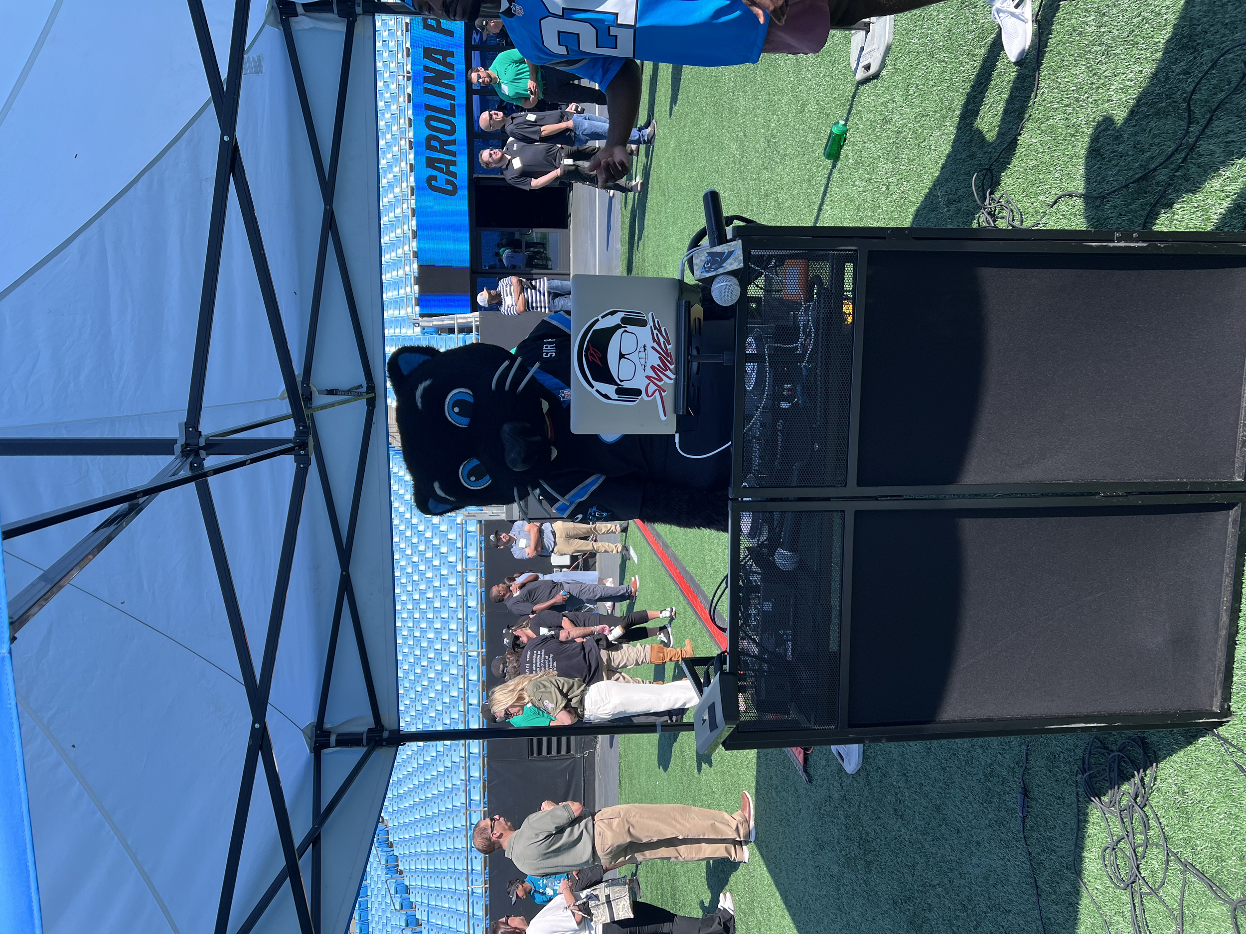 Panther mascot with a laptop on a table at a sports event, surrounded by people, on a green field with blue stadium seats in the background.