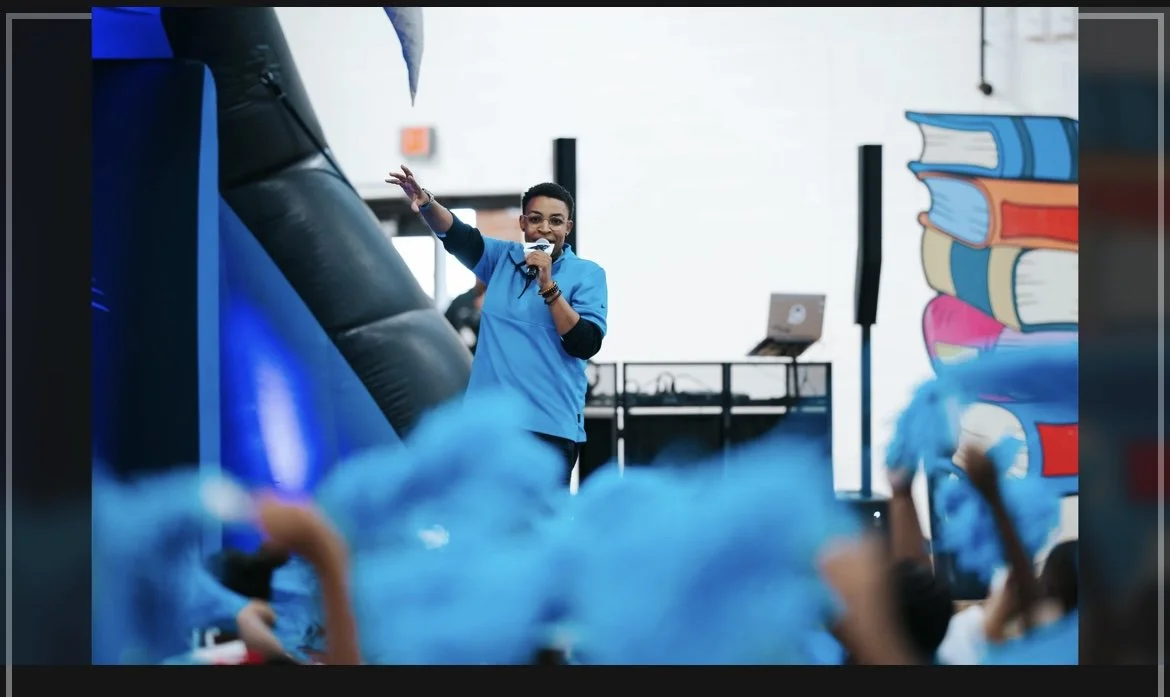 DJ Smylee speaking into a microphone at a school event with children dressed in blue in the foreground. There is a colorful cartoon book mural on the wall behind her.
