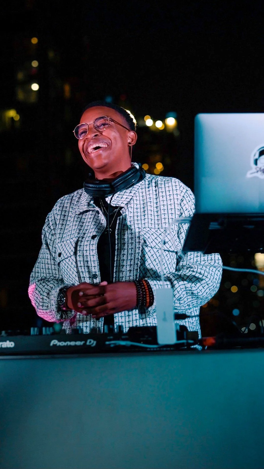 A joyful person with glasses and headphones around their neck, standing behind a DJ console with a laptop, at night in an outdoor setting with city lights