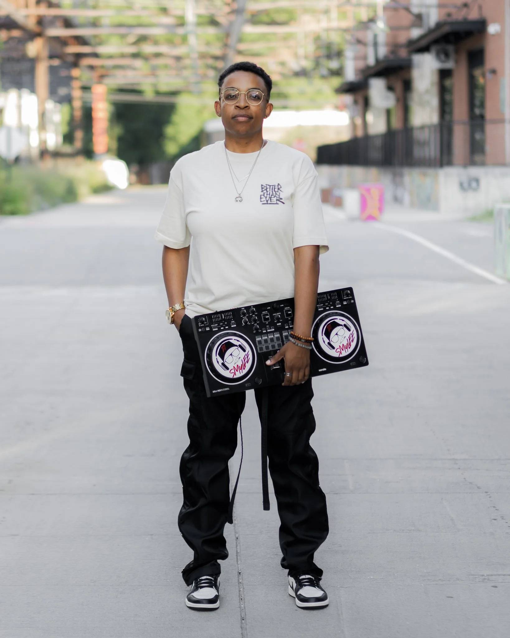 A person standing outdoors on a city street holding a DJ controller, wearing glasses, a white T-shirt, and black pants.