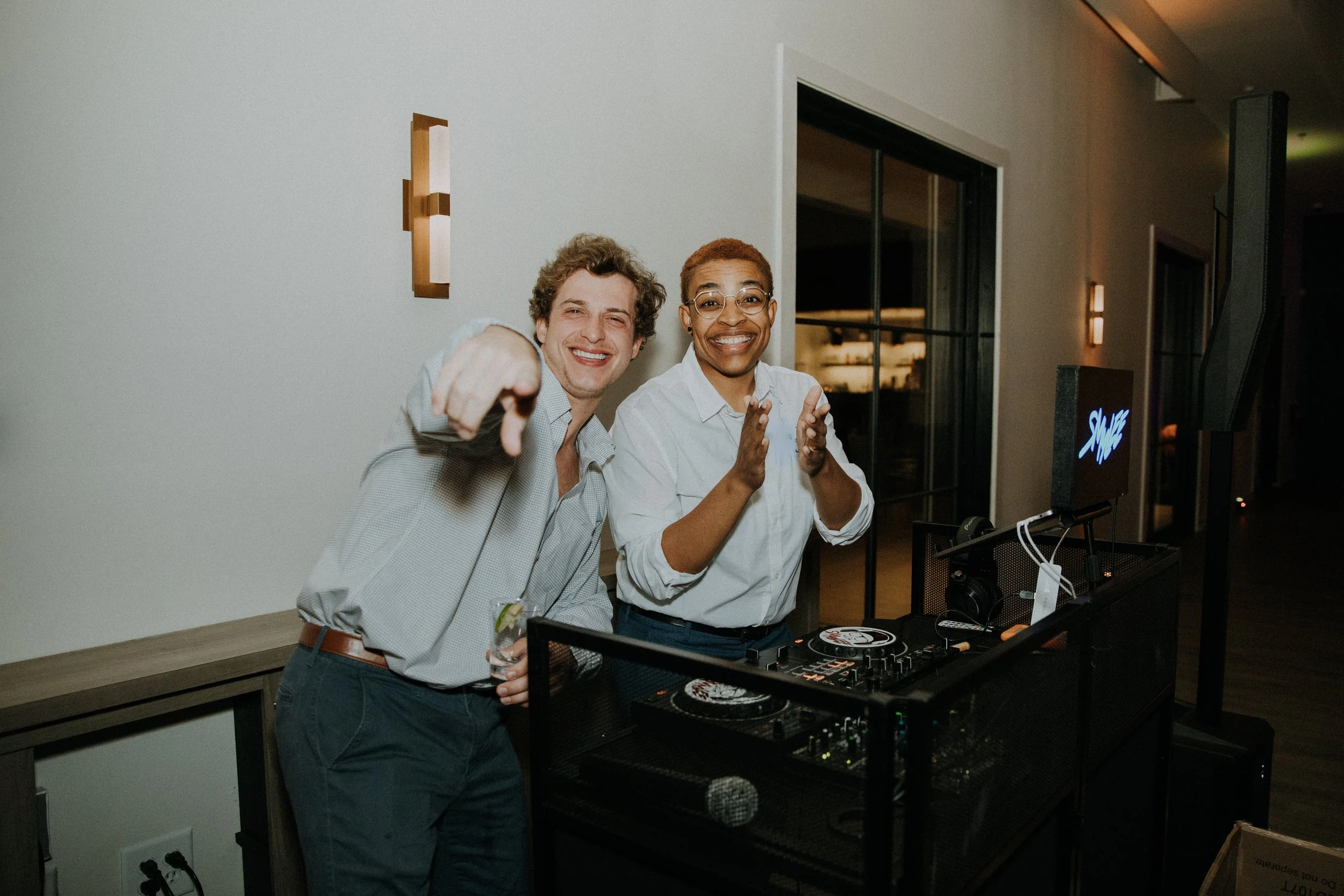 Two smiling people, a man and a woman, at a DJ booth or sound station in a celebration or event setting, with the man pointing at the camera.