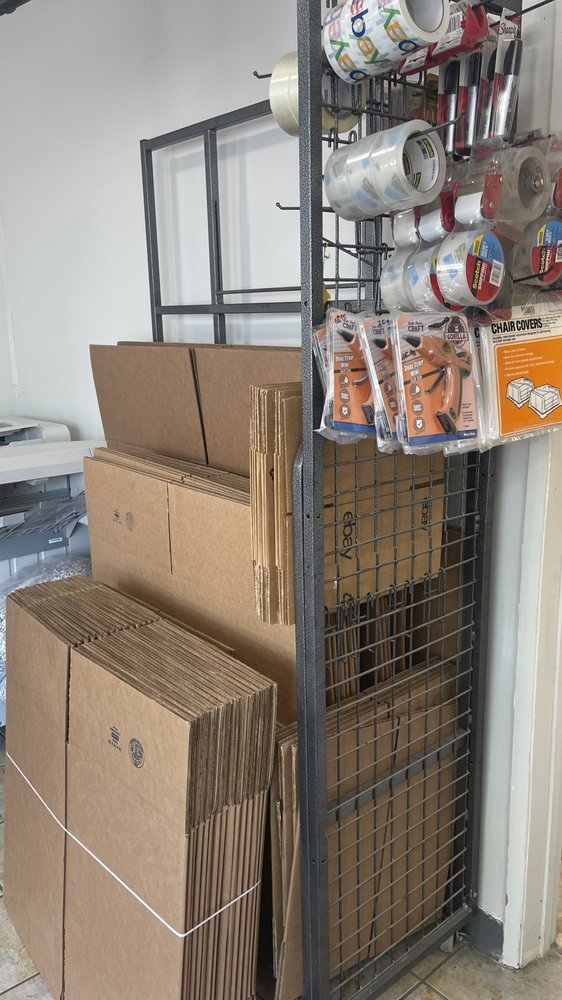 A metal shelving unit with boxes of empty cardboard and paperboard packaging, plastic tape, and various packing supplies, including scissors and chair covers.