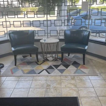Two black armchairs with a small round table between them, placed on a geometric-patterned rug near a large window with a decorative grid pattern.