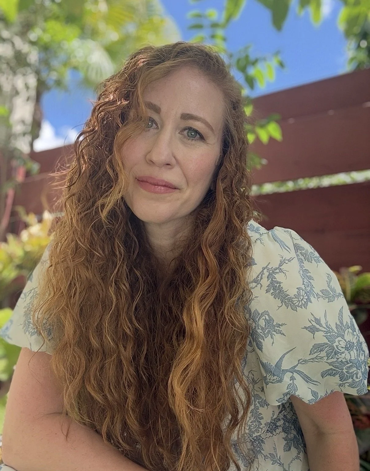 A woman with long, curly red hair and blue eyes poses outdoors on a sunny day, with a blue sky and green trees in the background.