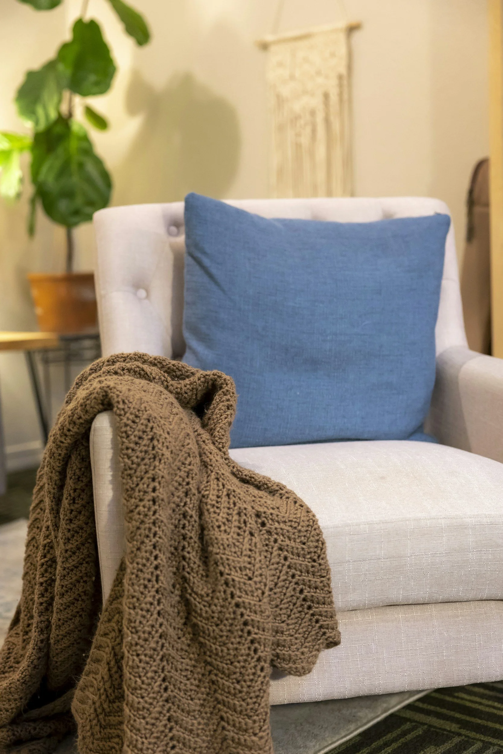 A cozy living room scene featuring a beige armchair with a large blue throw pillow. On the armchair, a chunky brown knitted blanket is draped. In the background, there is a large green potted plant and a woven wall hanging on a light-colored wall.