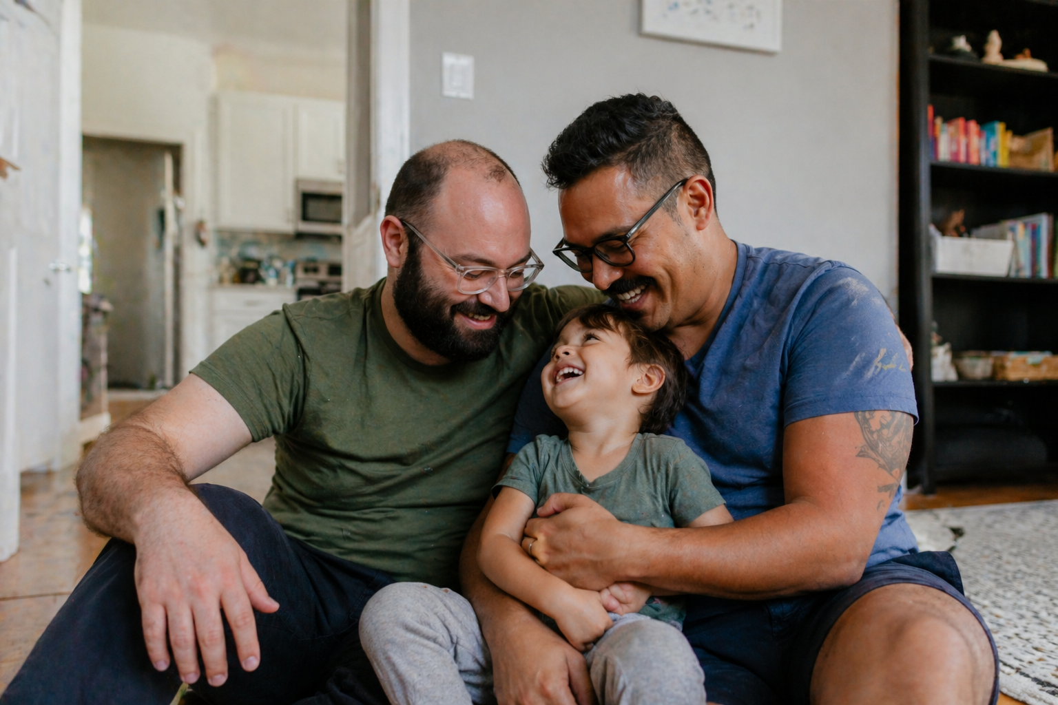 A happy family of two men and a young boy sitting on the floor indoors, smiling and embracing each other.
