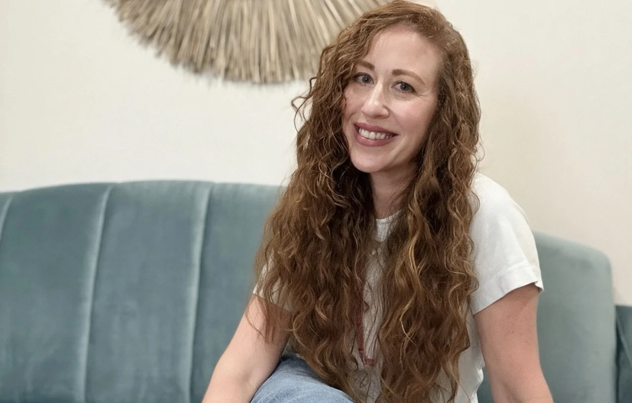 A woman with long, curly red hair sitting on a teal couch, smiling, in a room with a decorative wall hanging behind her.
