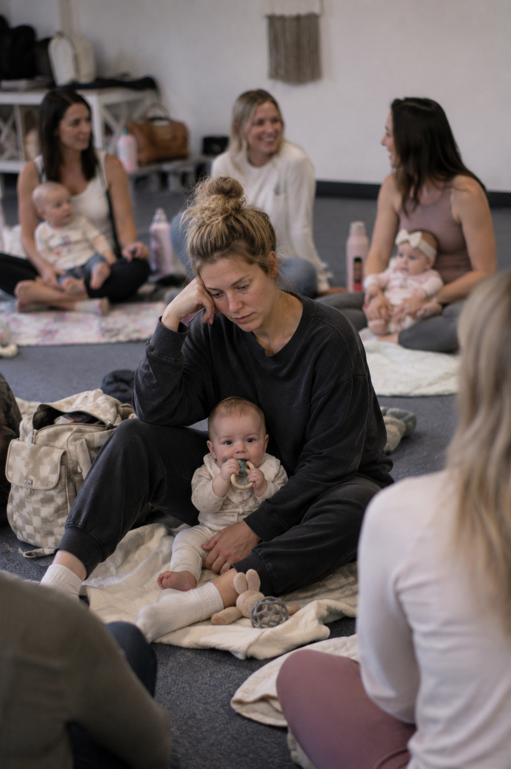 A group of women with babies sitting on the floor during a gathering, with one woman in the foreground appearing tired and holding a baby in her lap, while others chat and smile in the background.