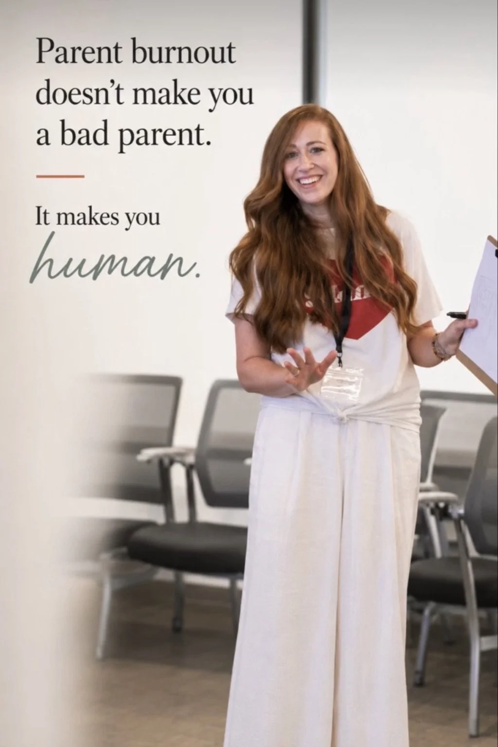 A smiling woman with long, red hair holding a clipboard and a pen, standing in front of a sign that reads "Parent burnout doesn't make you a bad parent. It makes you human." in a waiting room with black chairs.