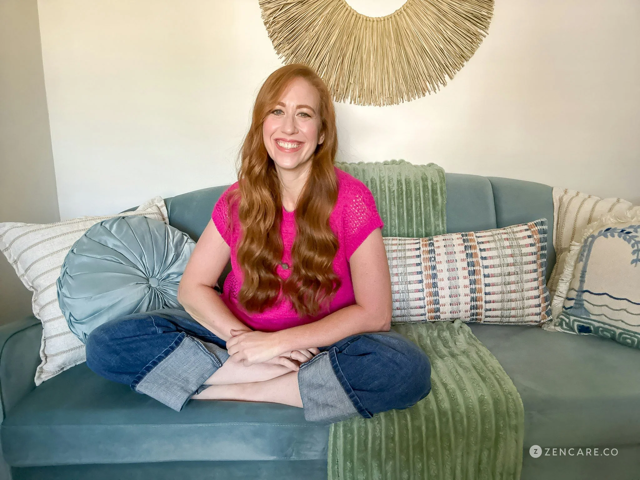 A woman with long red hair sitting cross-legged on a green couch, smiling, with various patterned pillows behind her and a decorative wall hanging above.