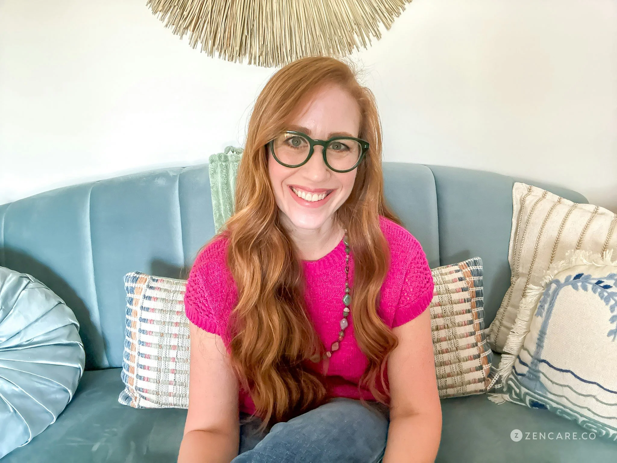 A woman with red hair, wearing glasses and a bright pink top, sitting on a couch surrounded by patterned cushions.