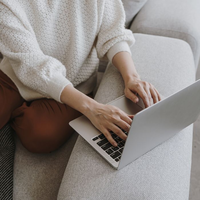 Person typing on a silver laptop while sitting on a gray sofa wearing a cream knitted sweater and brown pants.