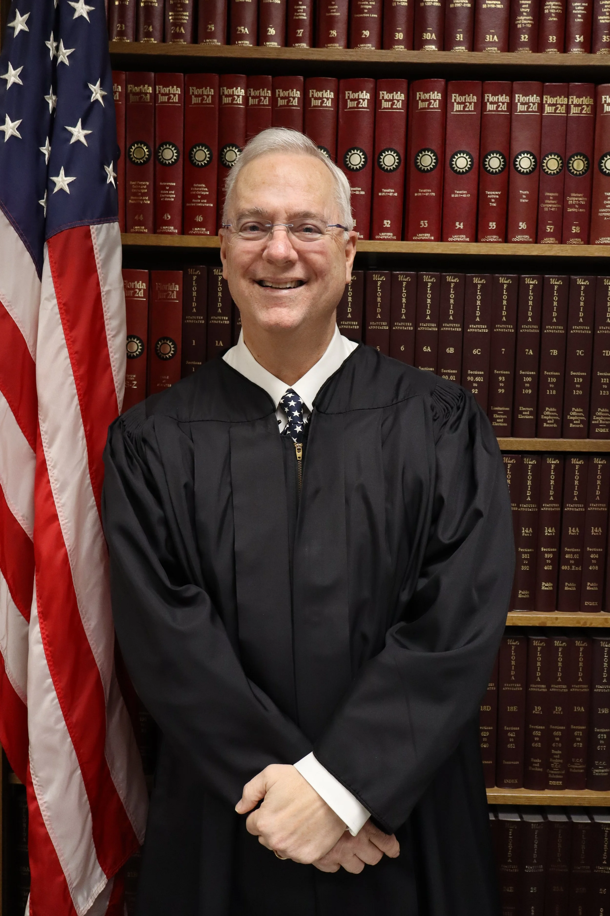 A man dressed in a black judicial robe with an American flag in the background, standing in front of legal books.