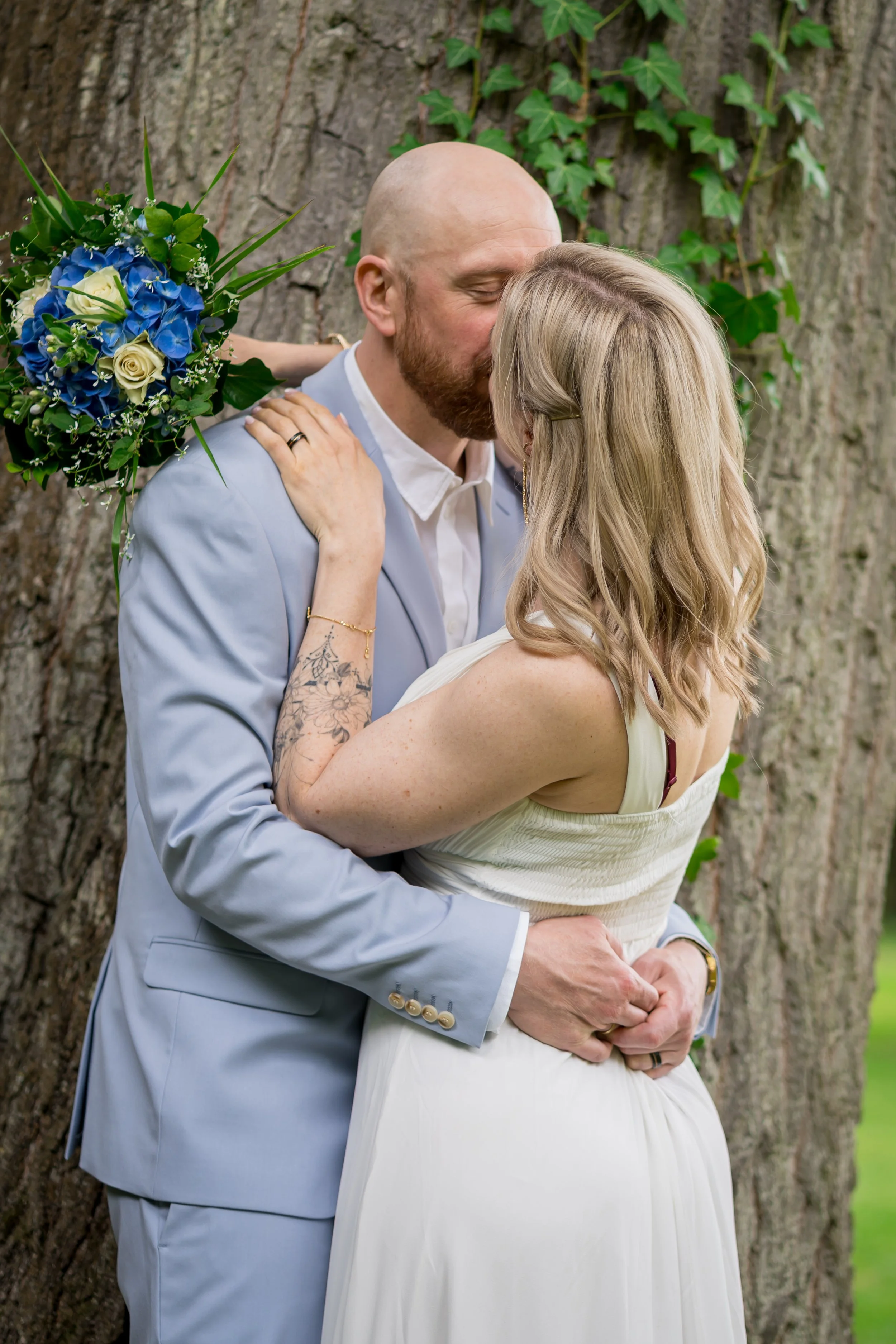 Paar beim Küssen im Freien, mit einer großen Baumstamm im Hintergrund und einer Frau, die einen blauen und weißen Blumenstrauß hält, bei einer Hochzeit oder romantischem Anlass.