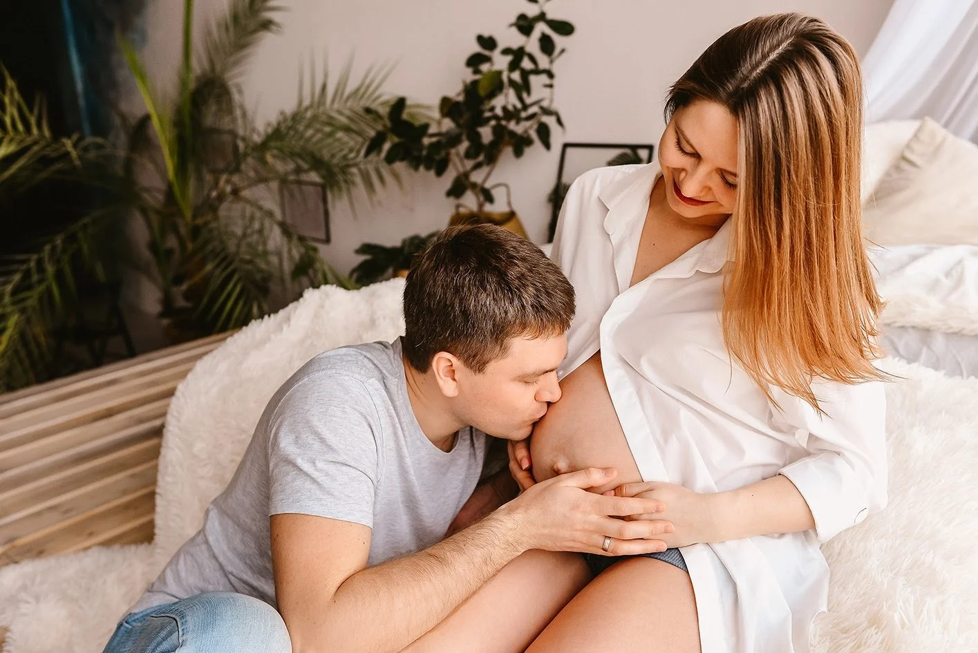 Ein Mann küsst während einer Schwangerschaft die schwangere Bauch einer Frau, die lachend neben ihm sitzt in einem gemütlichen Raum mit Pflanzen im Hintergrund.