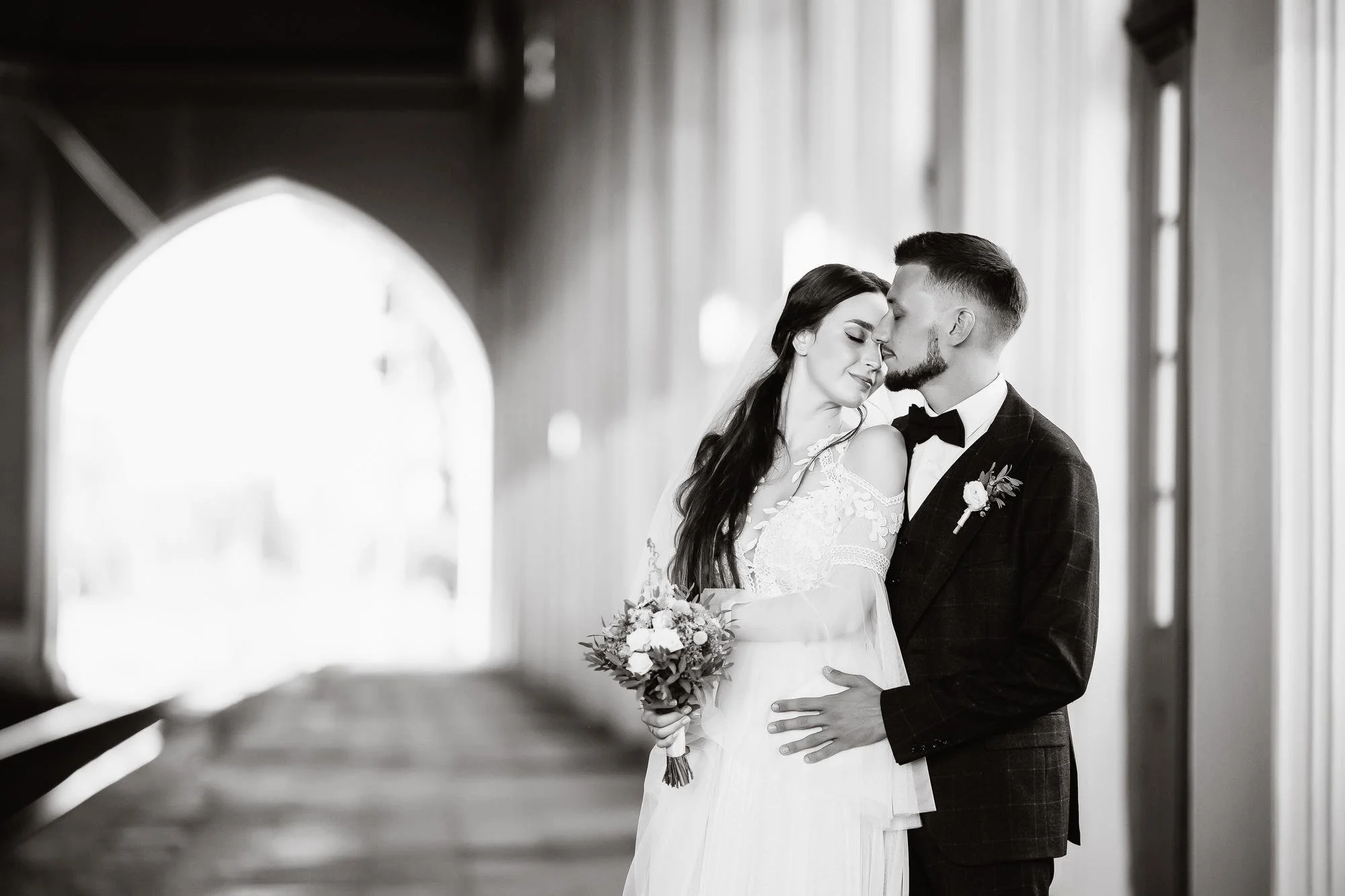 Braut und Bräutigam umarmen sich liebevoll in einer Kirche, schwarz-weiß Fotografie. Die Braut hält einen Blumenstrauß, beide tragen formelle Hochzeitskleidung.