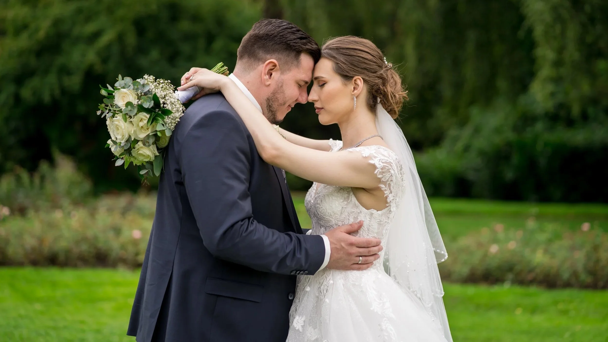 Ein Brautpaar steht in einem Garten und küsst sich zärtlich, die Braut trägt ein weißes Hochzeitskleid mit Spitze, der Bräutigam einen dunklen Anzug, sie umarmen sich liebevoll.