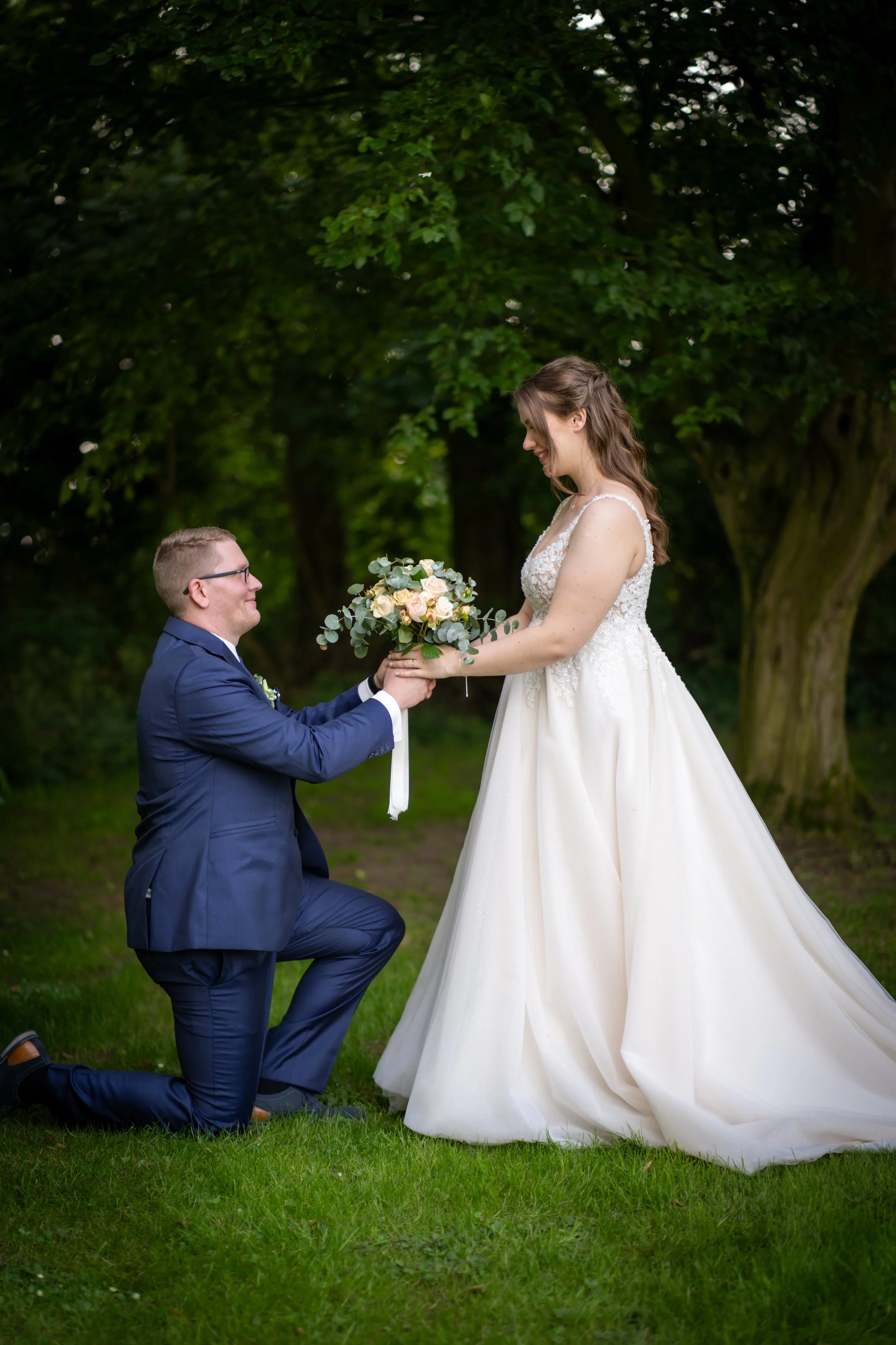 Ein Mann kniet im grauen Anzug auf dem Rasen, hält die Hand einer braunhaarigen Frau in einem weißen Brautkleid, während er ihr einen Blumenstrauß überreicht, im Hintergrund Bäume im Park.