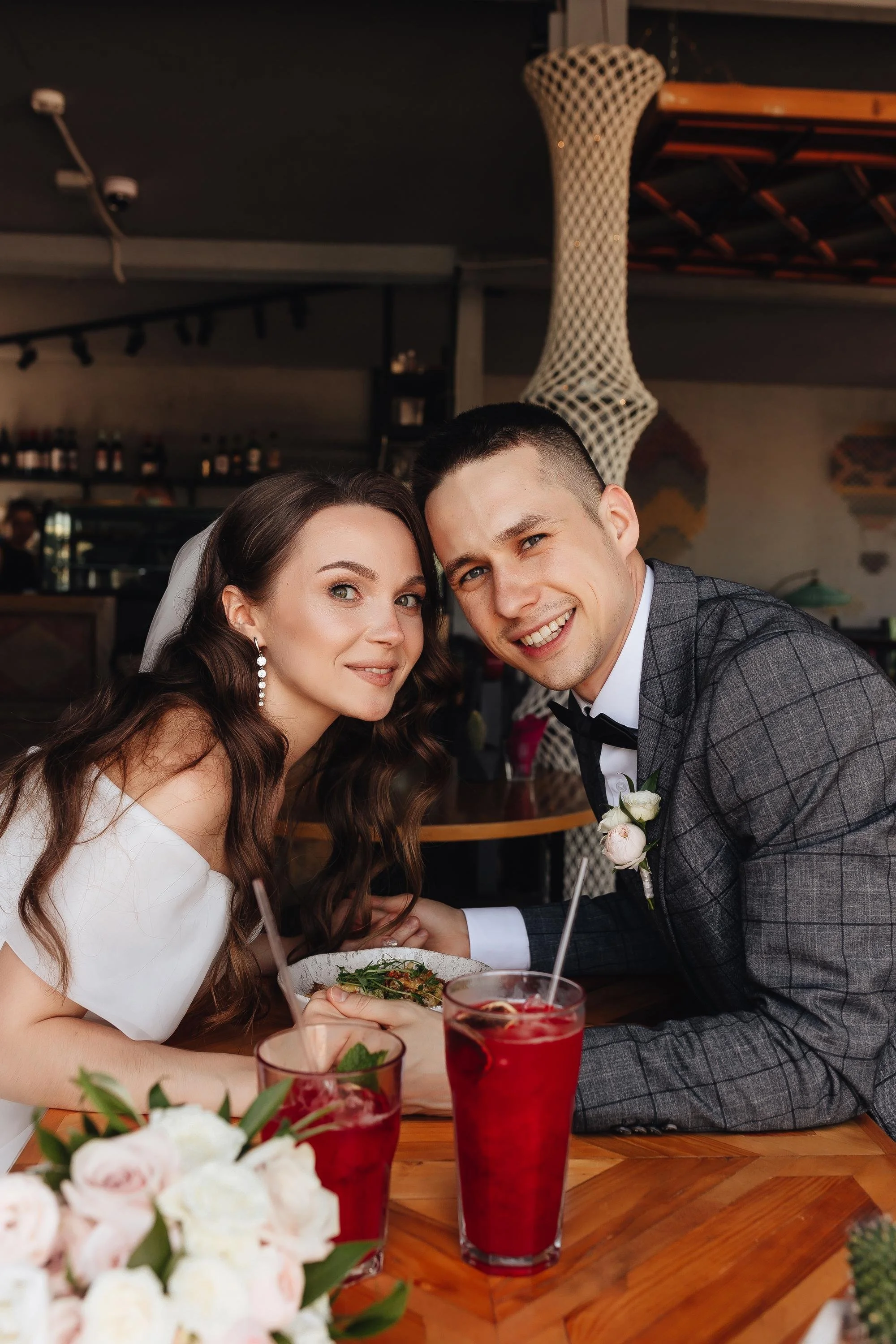 Ein glückliches Brautpaar sitzt bei einem festlichen Anlass in einem Restaurant, umgeben von Blumen, mit zwei roten Getränken auf dem Tisch.