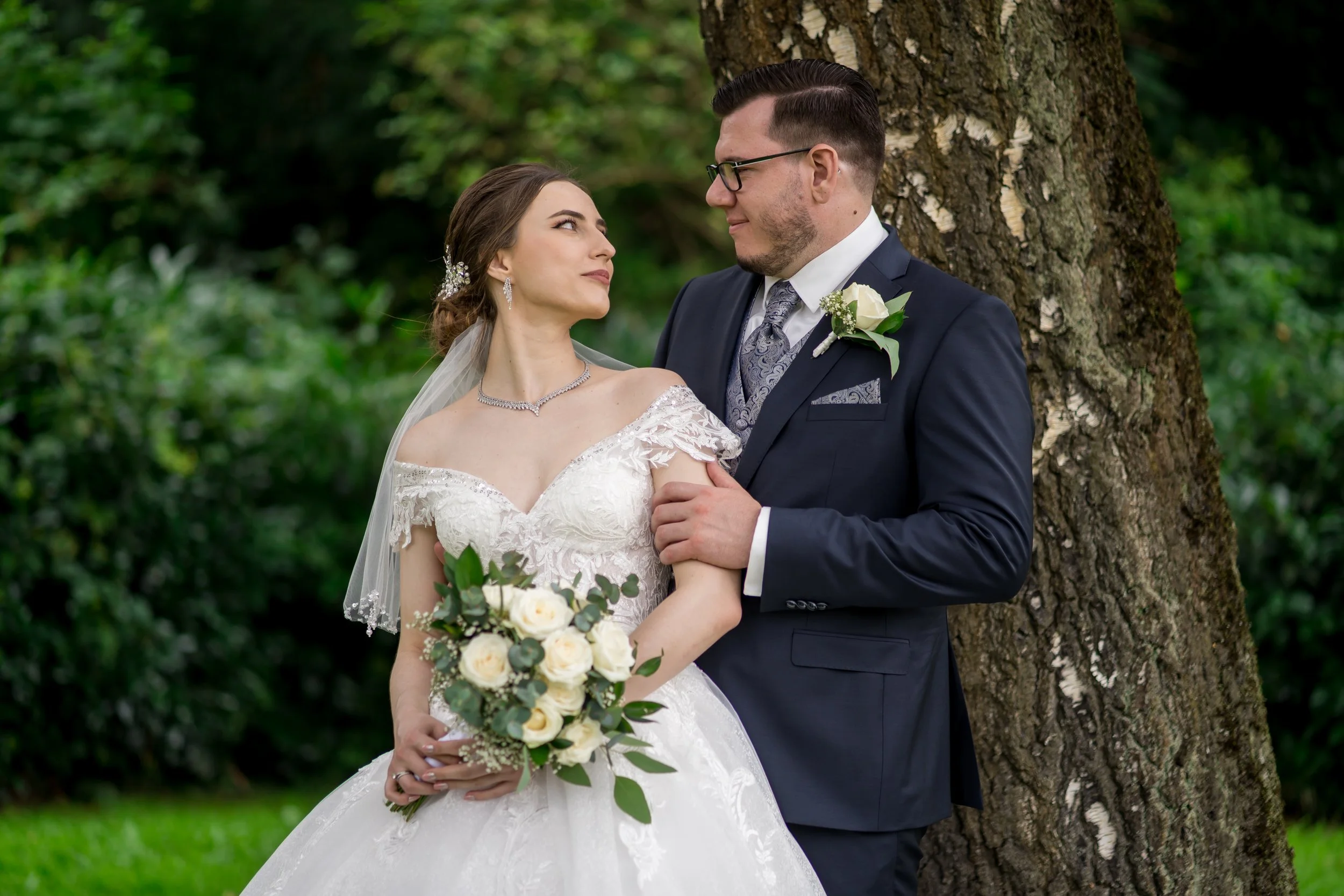 Ein brautpaar in formeller Kleidung steht im Grünen, die Braut hält einen Blumenstrauß, beide schauen sich verliebt an.