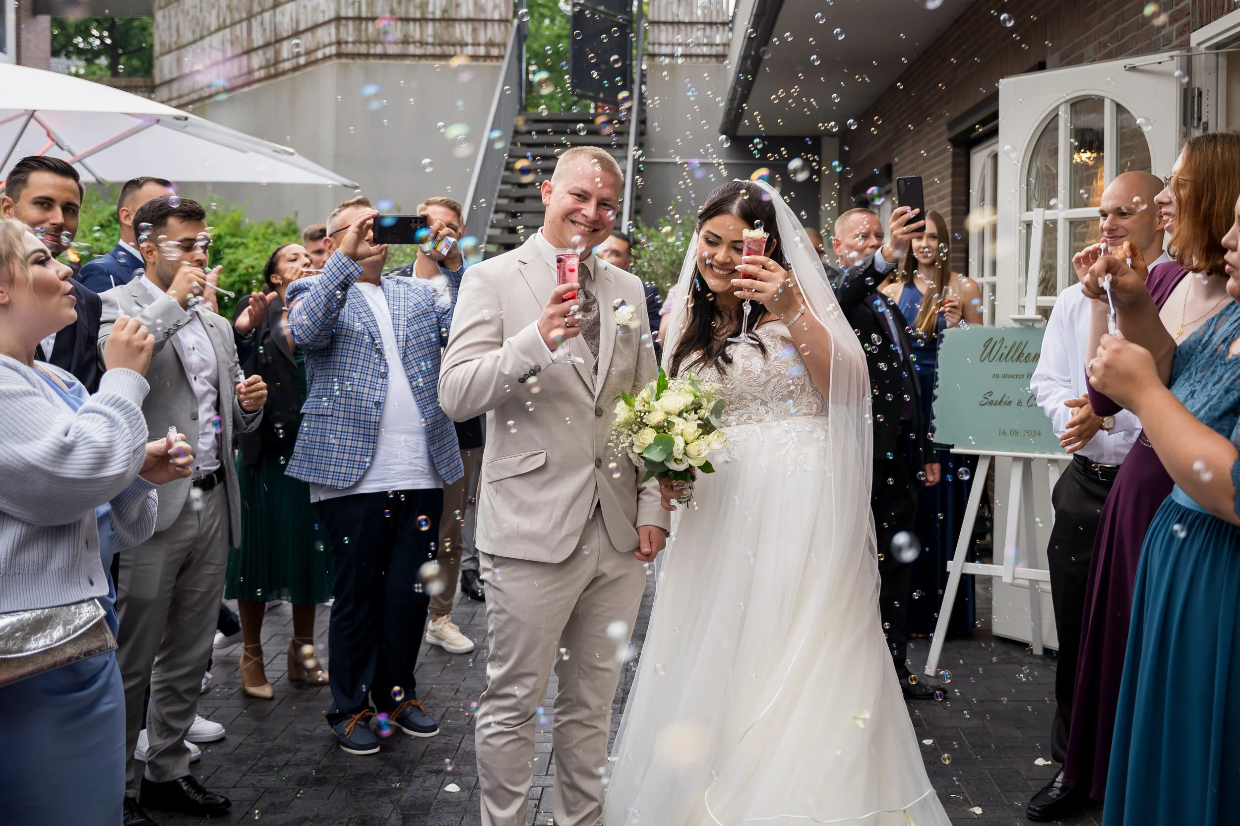 Hochzeitspaar umgeben von Freunden und Familie, die mit Konfetti und Seifenblasen feiern, draußen vor einem Haus.