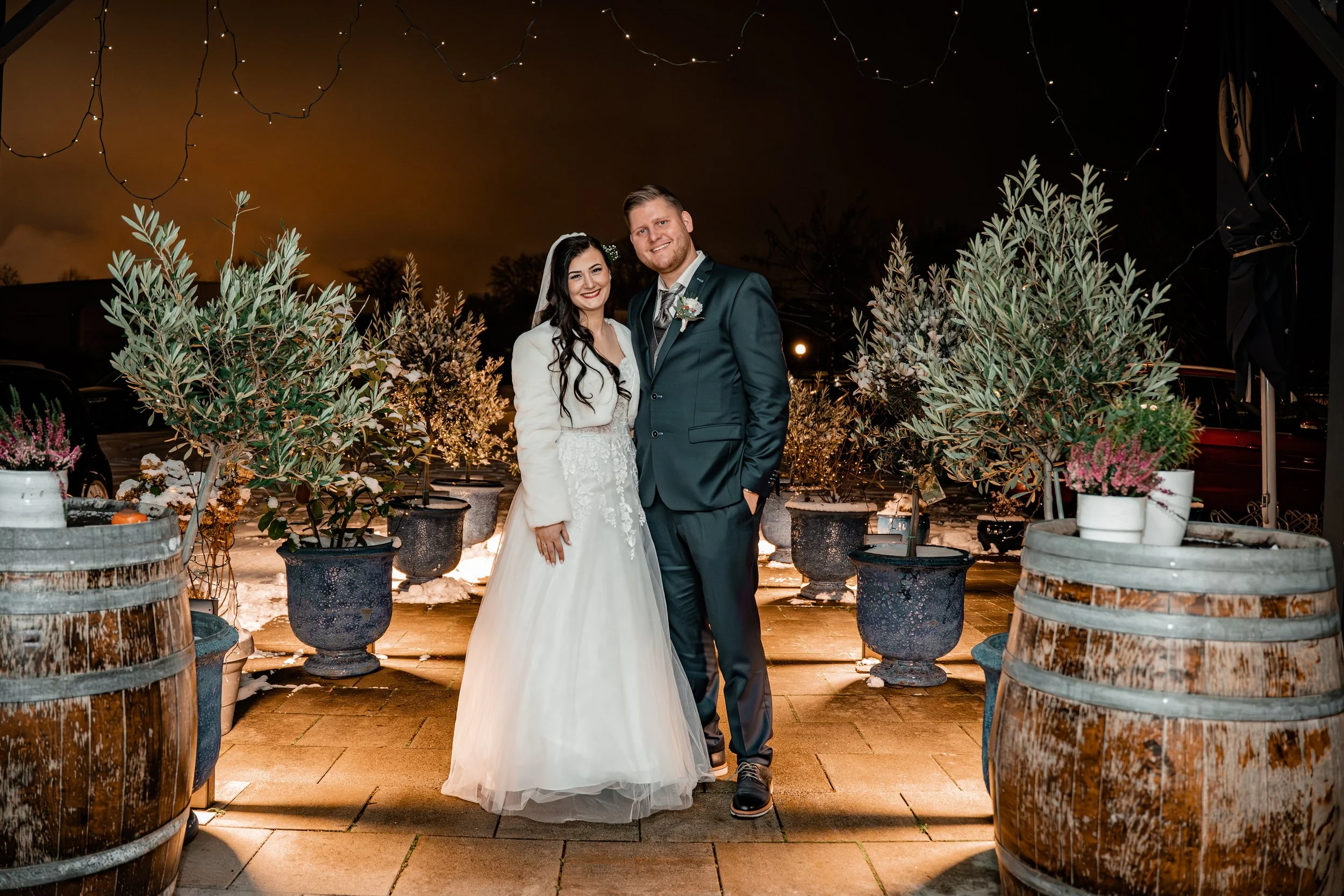 Ein junges Brautpaar bei Nacht, umgeben von Pflanzen in Töpfen und Holzfässern, bei einer Winterhochzeit, mit Lichterketten im Hintergrund.