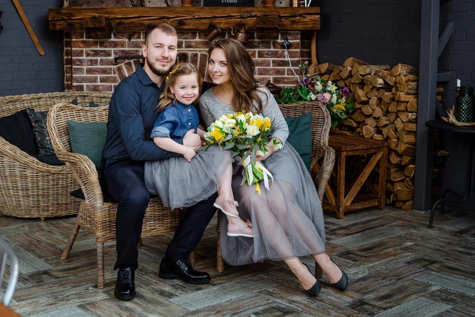 Familie auf einem Wicker-Sofa, das kleine Mädchen hält einen Blumenstrauß und sitzt auf dem Schoß der Mutter, während der Vater das Mädchen umarmt. Alle lächeln. Im Hintergrund ein Kamin aus Ziegeln, Brennholz und Blumenarrangements.