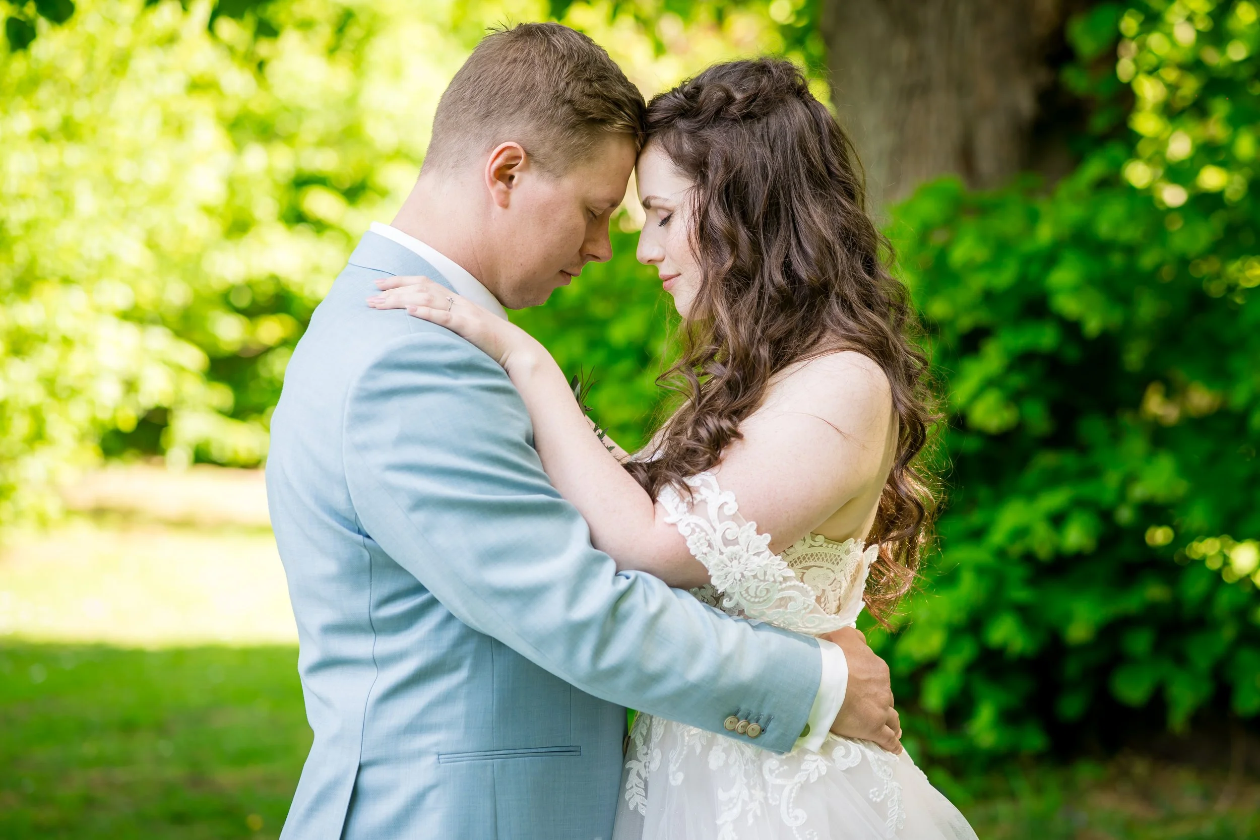 Ein Brautpaar steht sich in einem Park mit grünen Bäumen gegenüber, beide mit geschlossenen Augen, umarmt und zeigt Intimität und Liebe.