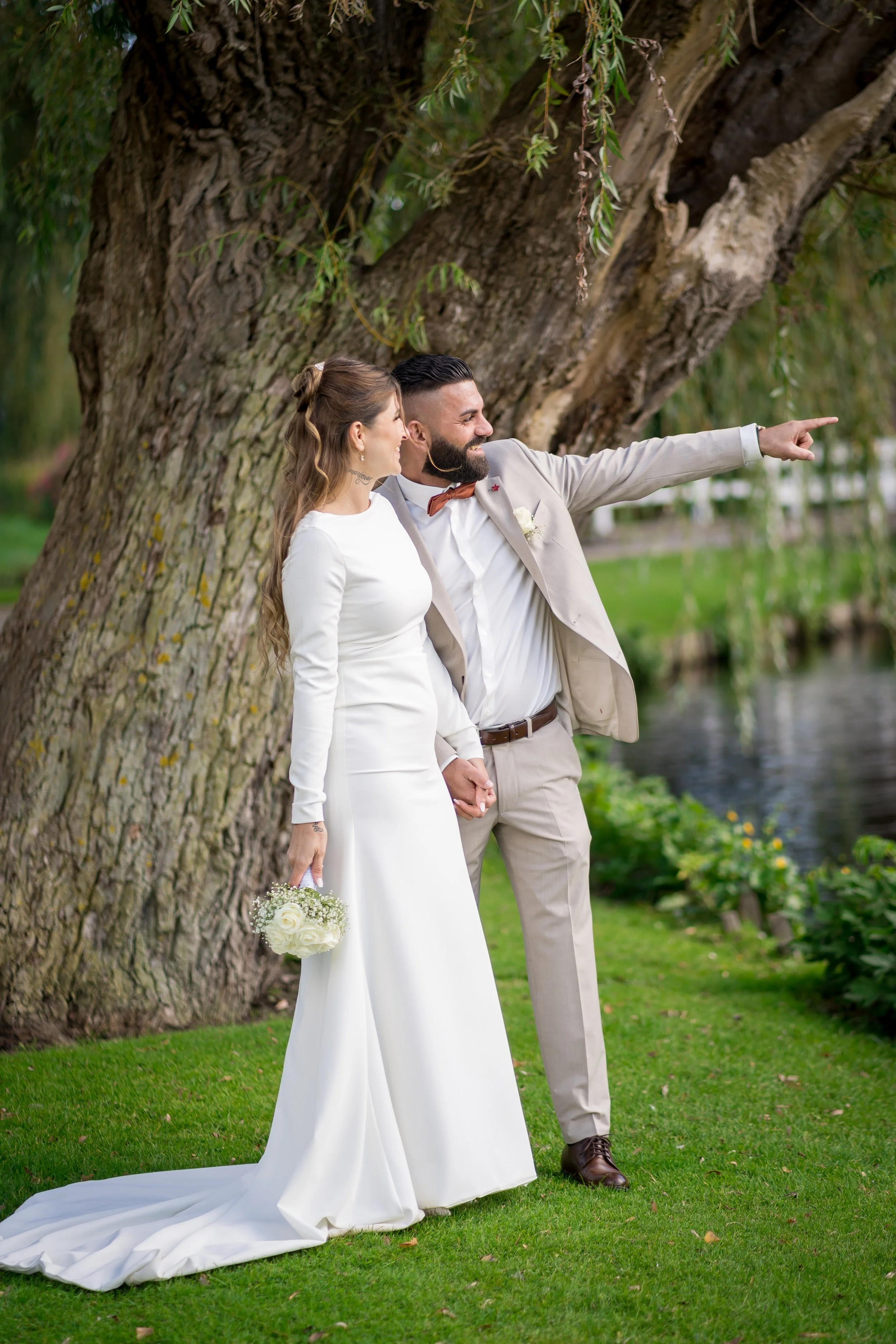 Ein Brautpaar steht am Ufer eines Flusses, die Frau in einem weißen Brautkleid hält einen Blumenstrauß und der Mann in einem hellen Anzug zeigt nach vorne, während sie sich an den Händen halten.