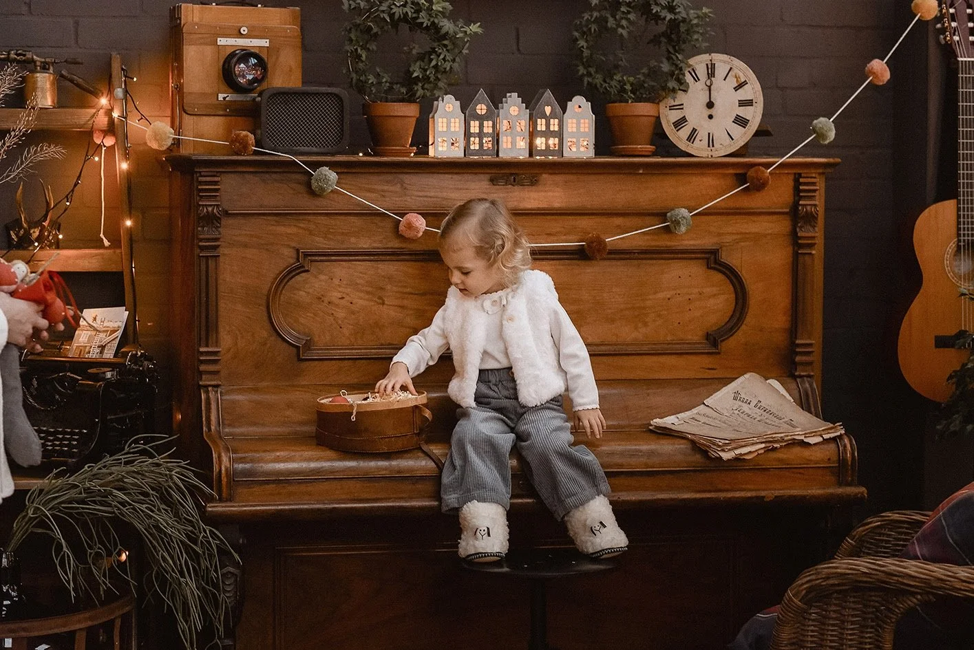Ein kleines Kind sitzt auf einem Hockstuhl vor einem alten Holzklavier, das mit Dekorationen wie Lampen, kleinen Häusern, Töpfen und einer Uhr geschmückt ist. Das Kind spielt mit einem runden Holzbehälter und trägt warme Kleidung.