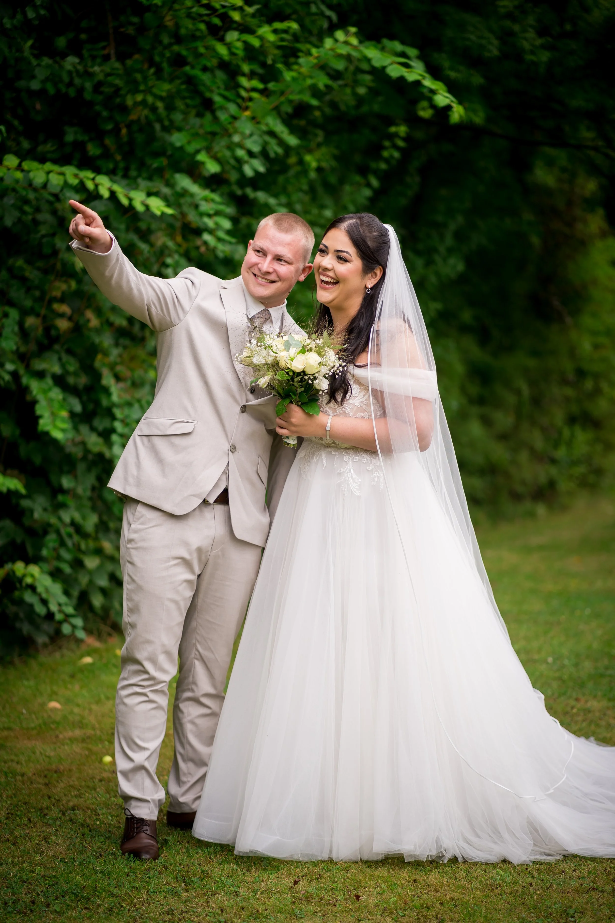 Braut und Bräutigam stehen im Garten, der Bräutigam zeigt auf etwas, beide lachen und halten einen Brautstrauß, umgeben von grünen Bäumen.