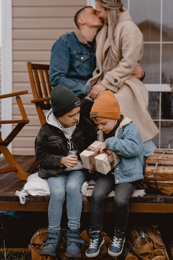 Eine Familie auf einer Veranda: Zwei Kinder sitzen auf einem Holzbank, eines hält ein Geschenk, während Mutter und Vater im Hintergrund sich küssen.