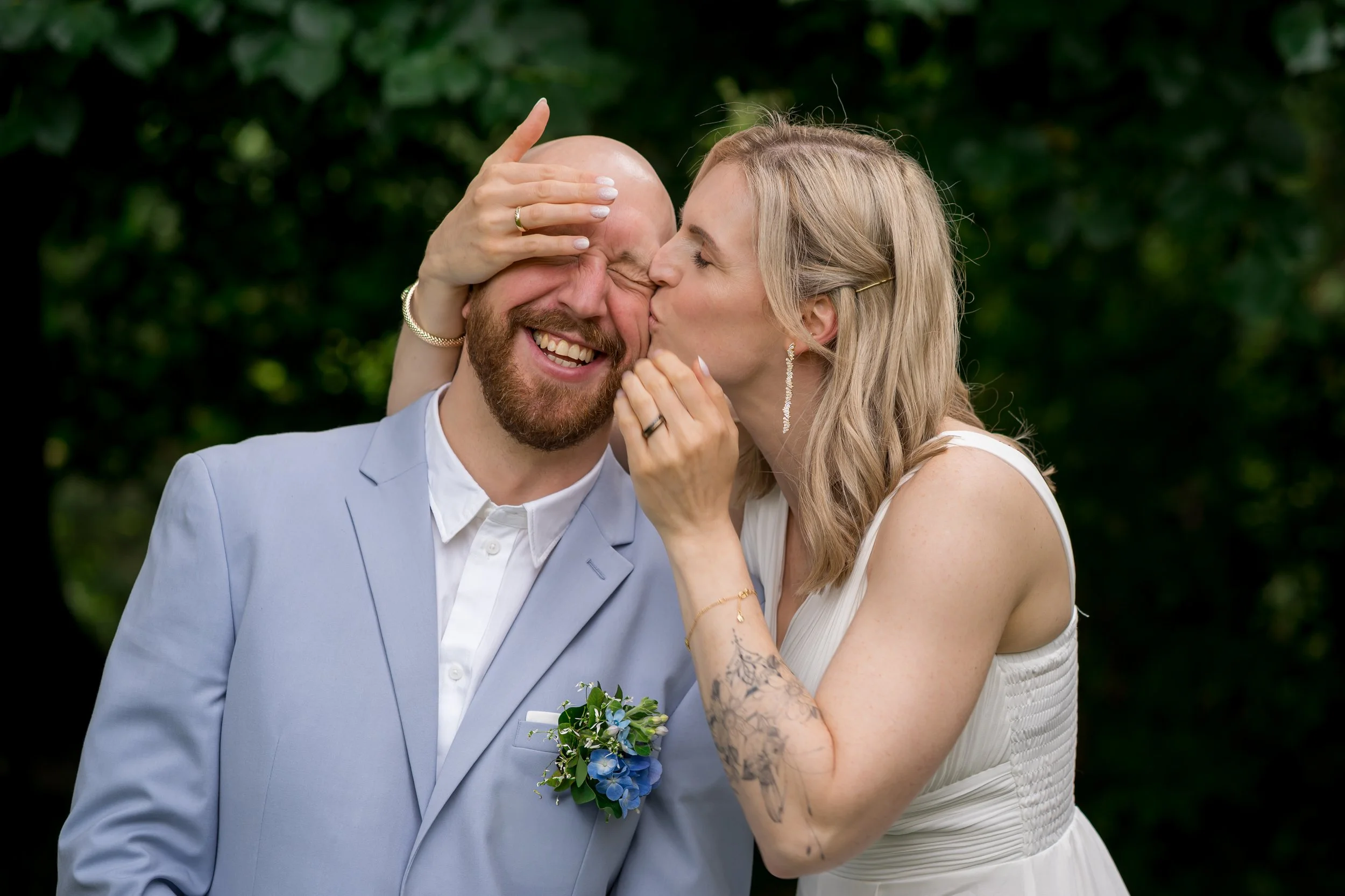 Eine Frau küsst einen lachenden Mann auf die Wange, während sie ihn liebevoll berührt, im Grünen.