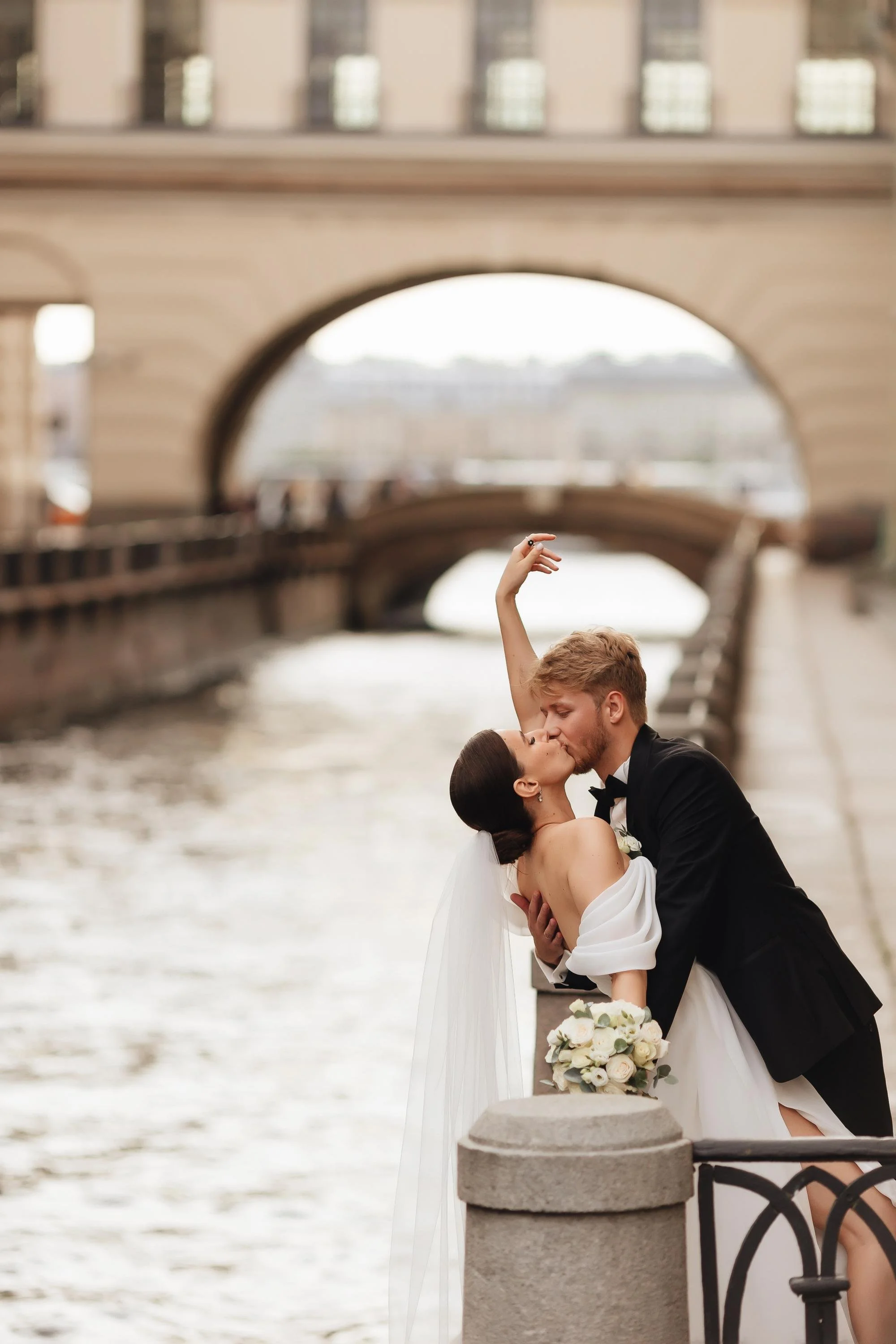 Ein frisch vermähltes Paar küsst sich an einem Flussufer unter einer Brücke, die im Hintergrund sichtbar ist, mit einer Braut in Weiß und einem Bräutigam in einem schwarzen Anzug.