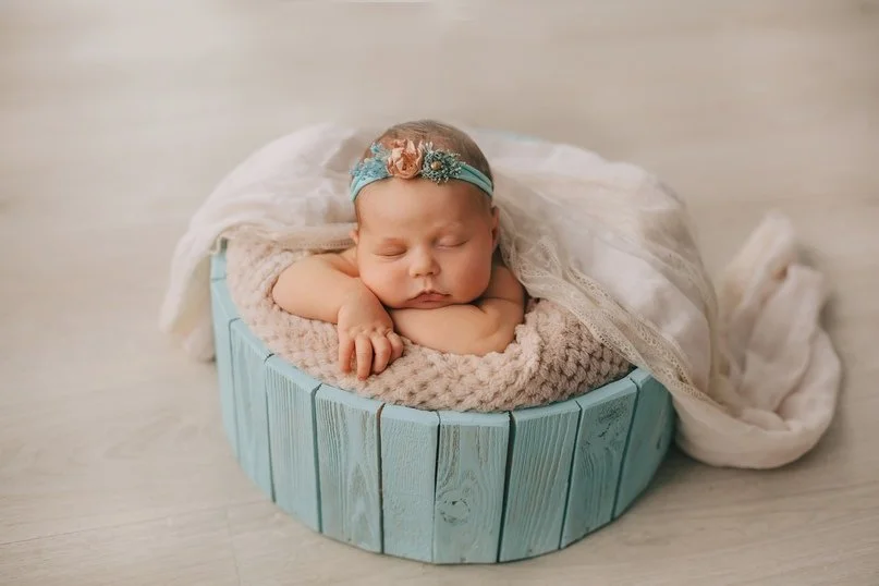 Ein schlafendes Baby mit Blumenhaarschmuck, das in einer runden, blaue Holz-Box liegt, umgeben von weichem Tuch und Decke.