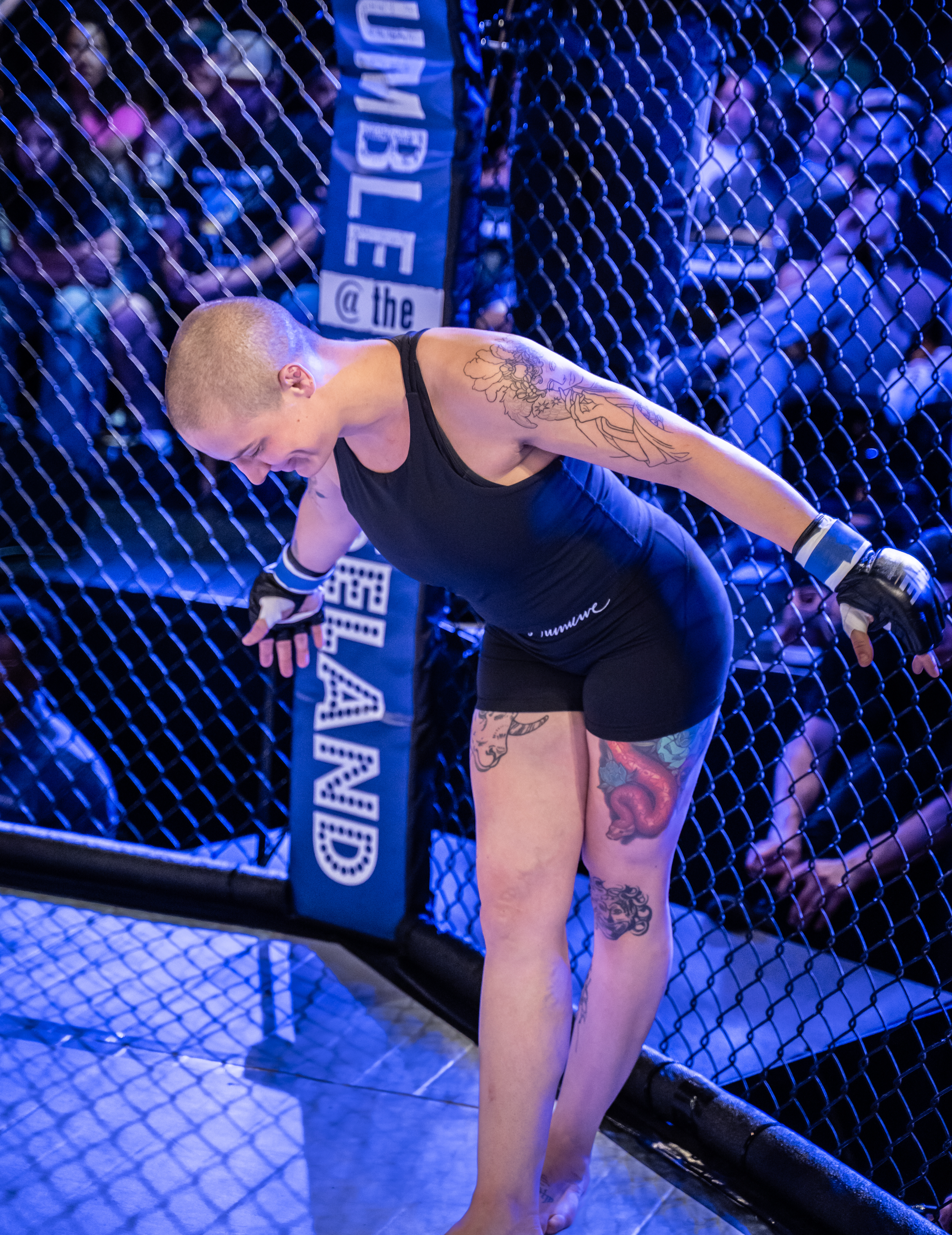 Female mixed martial artist with tattoos, bent near the cage, wearing black shorts and gloves, inside a UFC octagon.