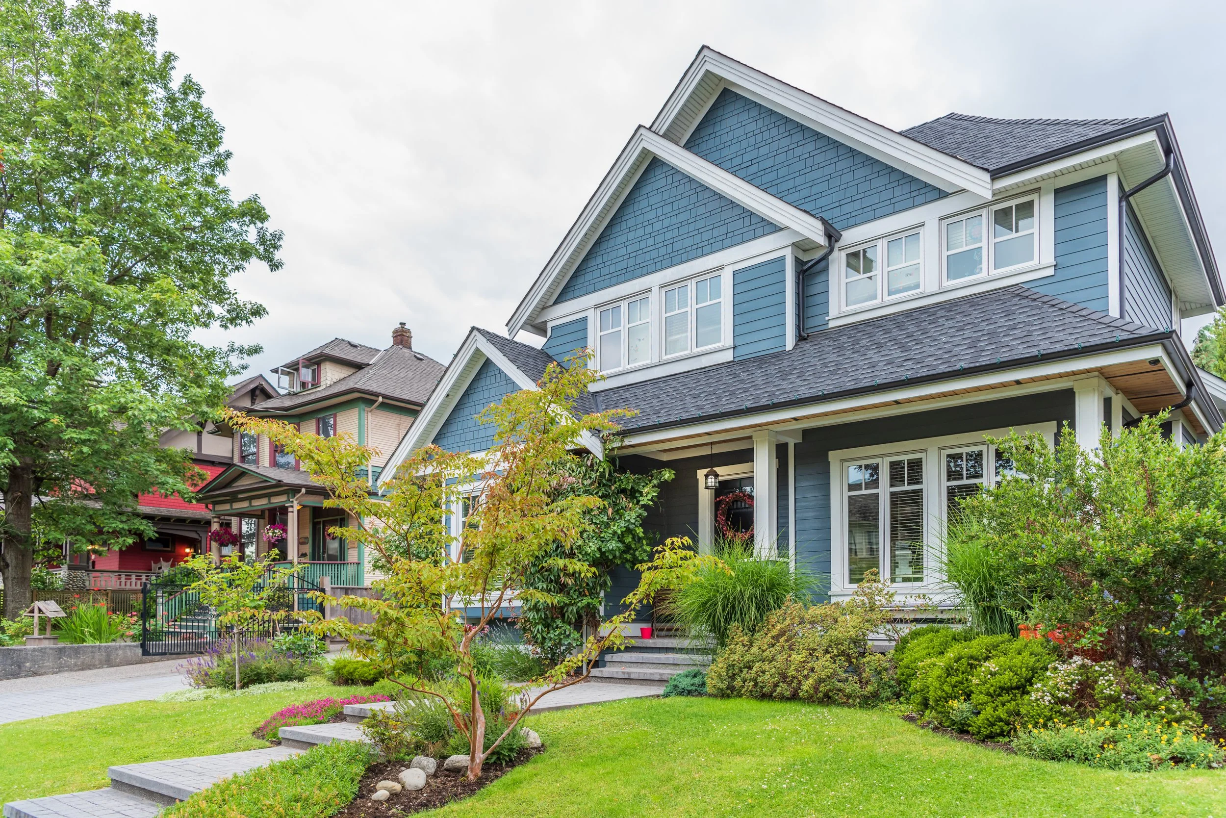 A two-story blue house with white trim and a front porch, surrounded by a well-maintained garden with various shrubs and trees.