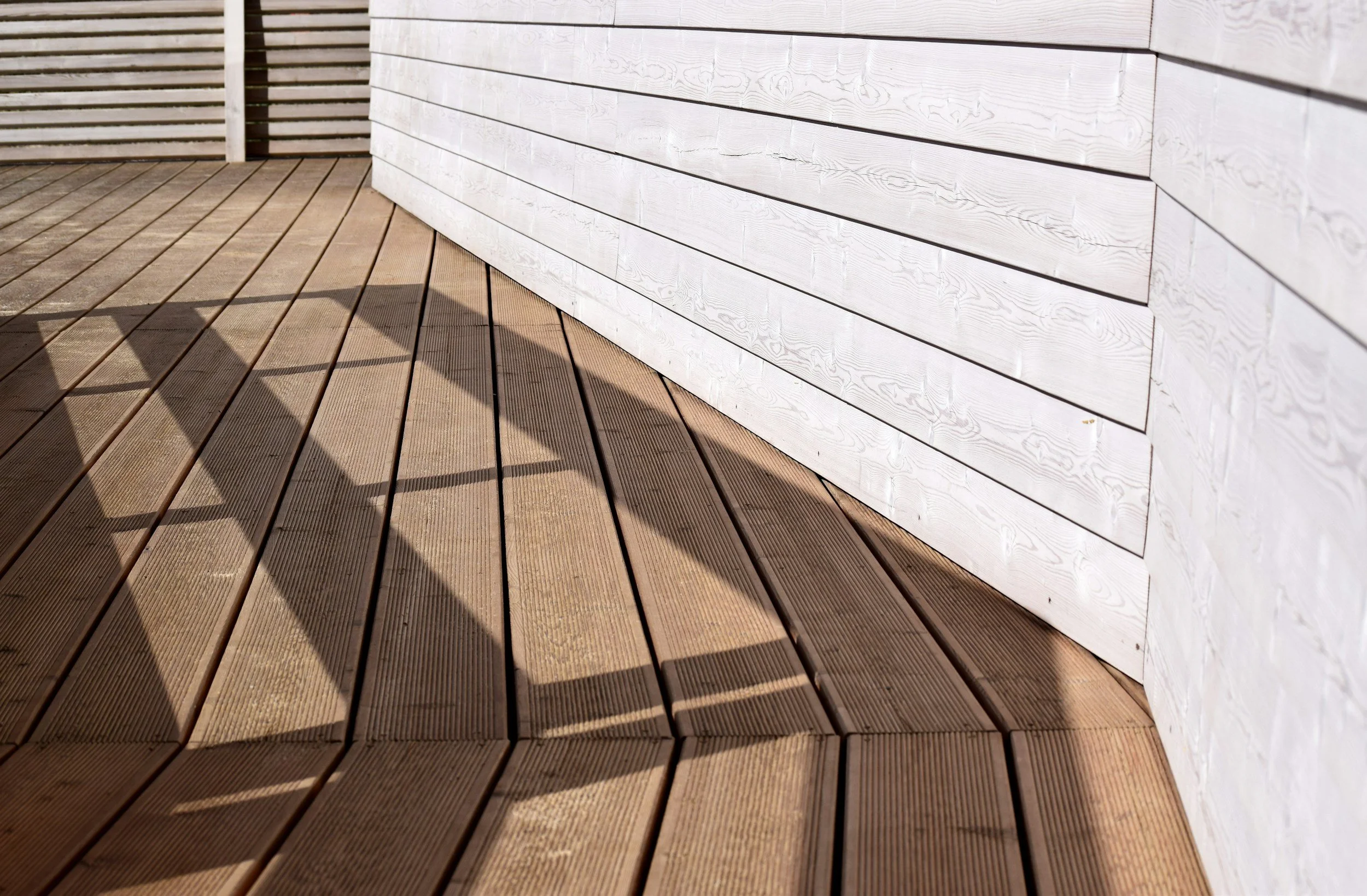 Close-up of a wooden deck with a shadow of a railing cast on it, and a white wooden wall with horizontal planks.
