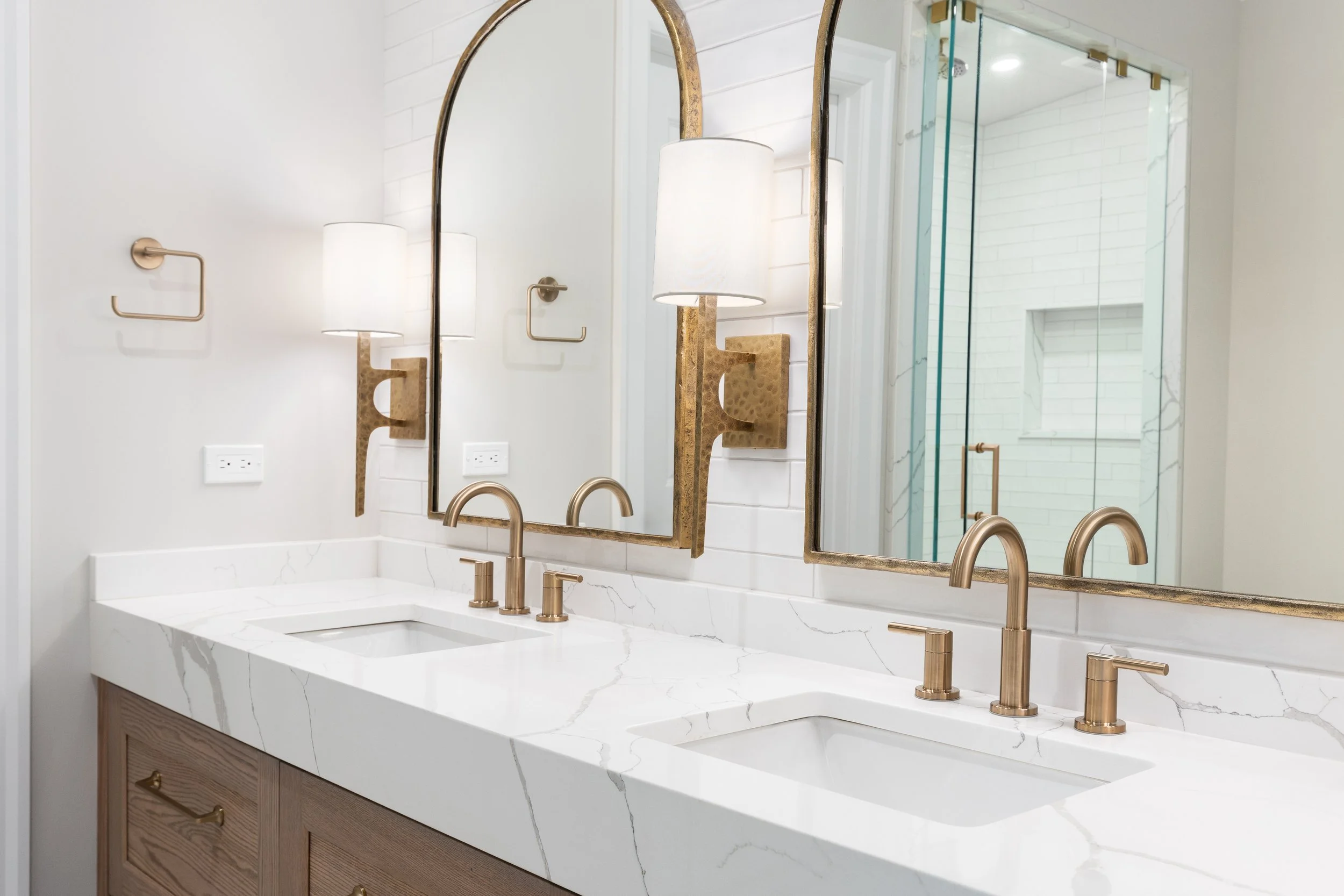 Double bathroom vanity with white marble countertop, two undermount sinks, brushed gold faucets, and two large mirrors. There are two wall sconces with white shades and brass accents, and a shower enclosure with glass doors and white brick walls visible in the background.