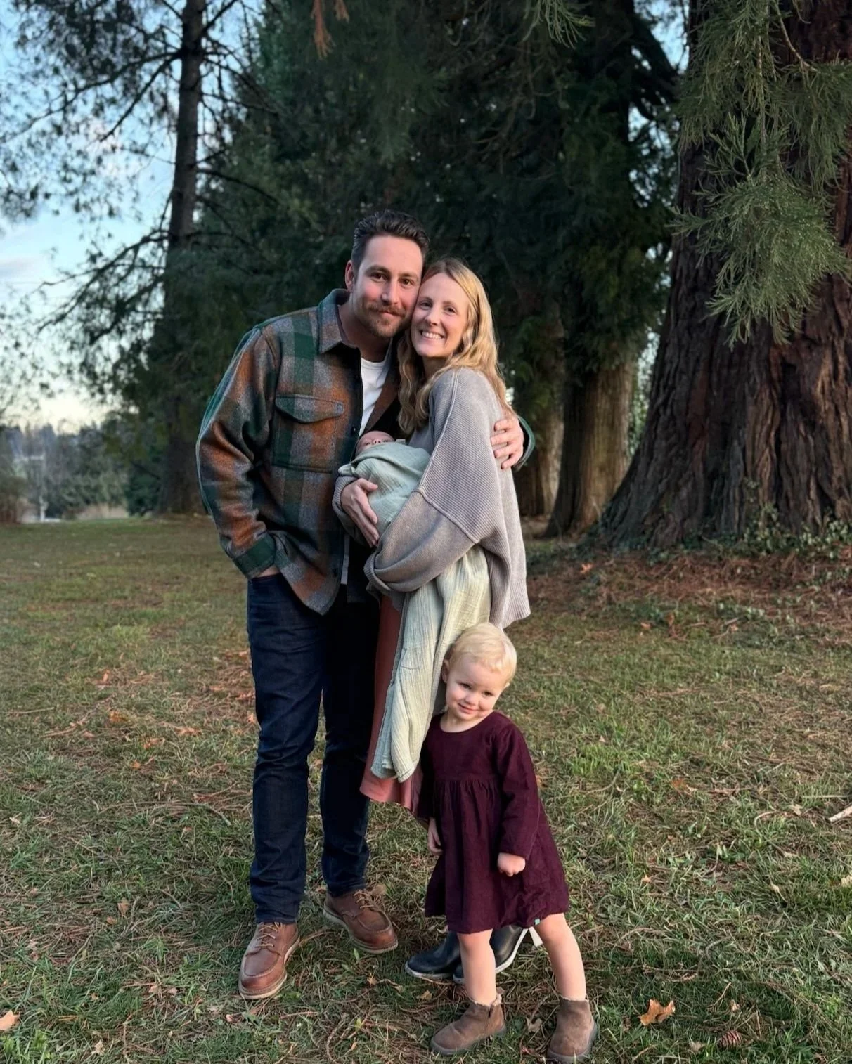 A family of four stands outdoors in a park with large trees in the background. The man and woman are smiling and hugging. The woman is carrying a baby wrapped in a blanket, and a young girl in a burgundy dress stands in front, leaning on one leg with a smile.
