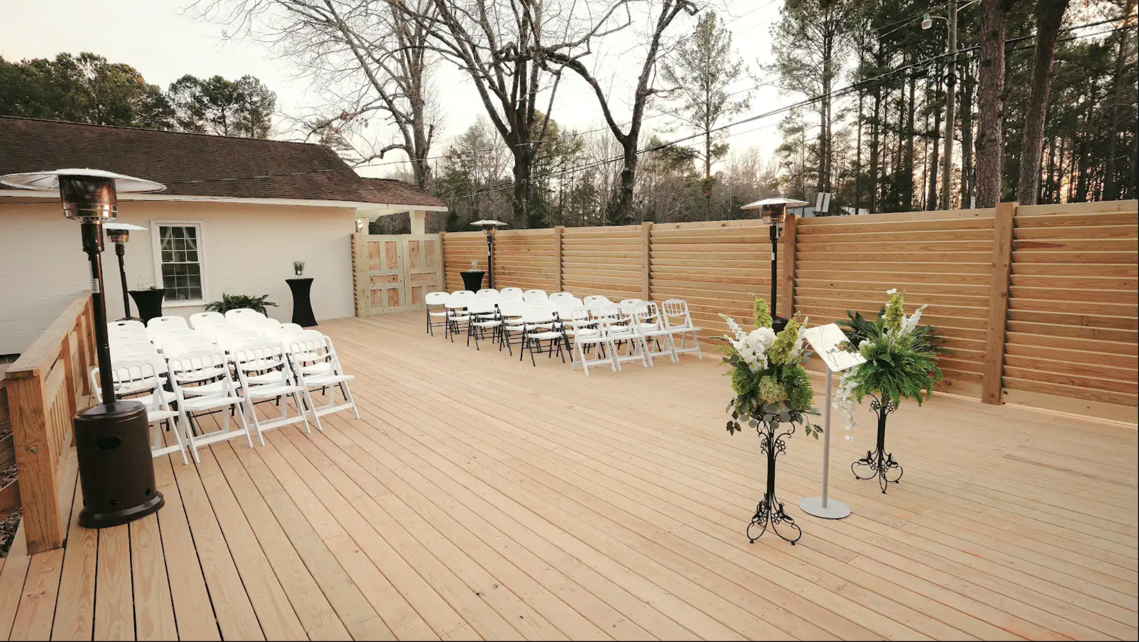 Empty wooden outdoor patio decorated for a wedding with two floral arrangements on stands and rows of white chairs facing a music stand.