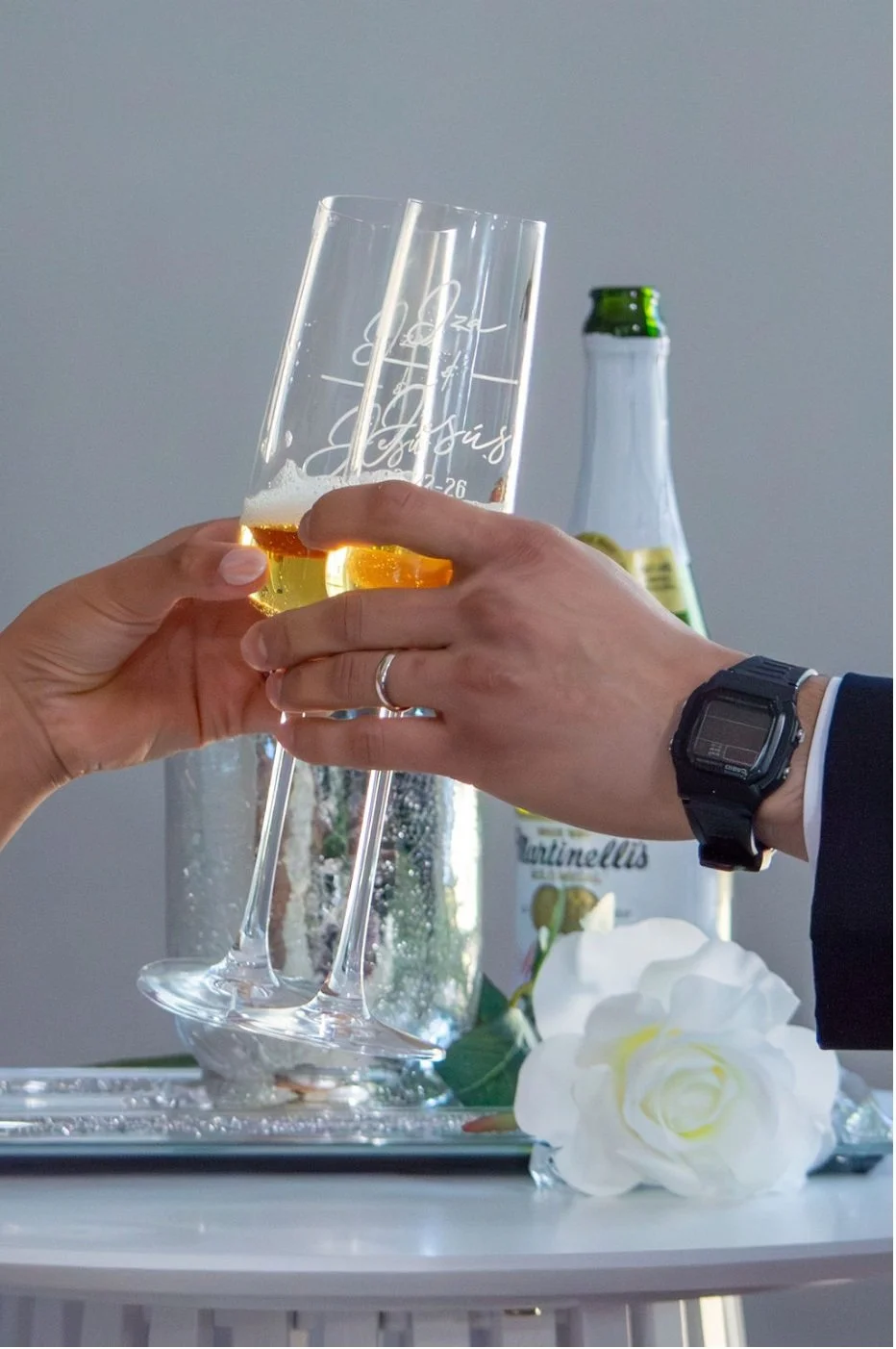 Two people toasting with champagne glasses, with a bottle of beer and white flowers on a table, celebrating a special occasion.