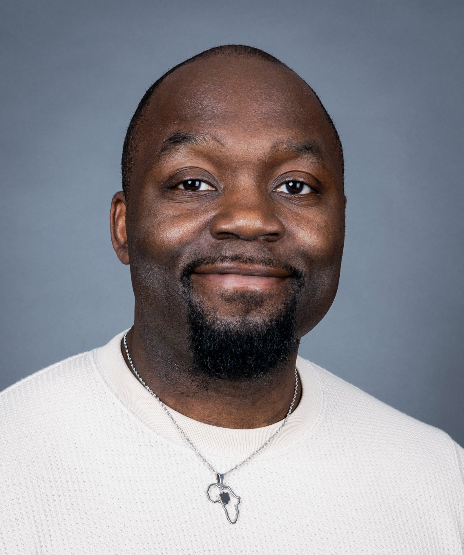 Headshot of a smiling Black man with a short beard and shaved head, wearing a light-colored shirt and a necklace with an Africa-shaped pendant, against a gray background.